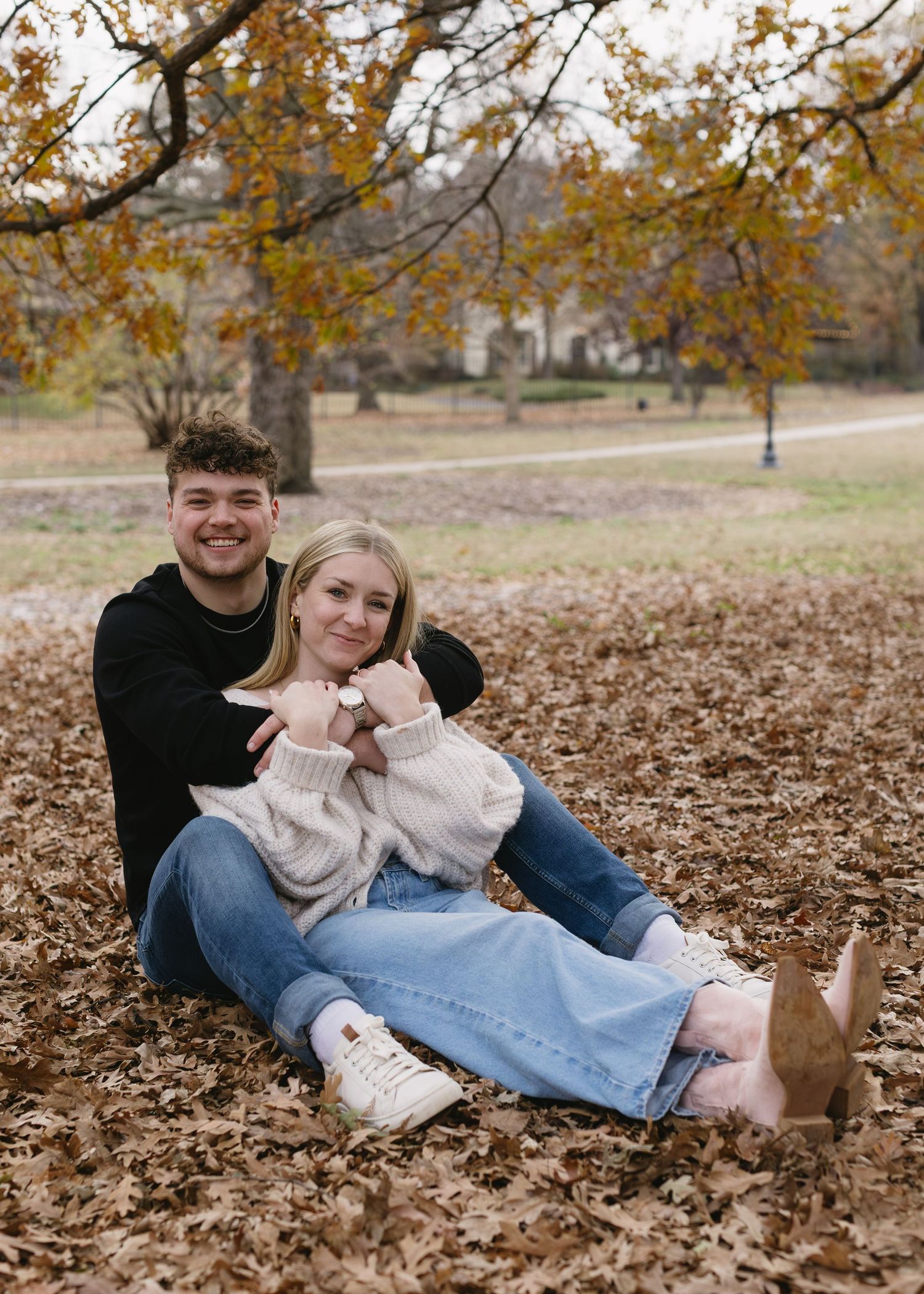 A couple sitting together on fallen autumn leaves making heart shapes with their hands.