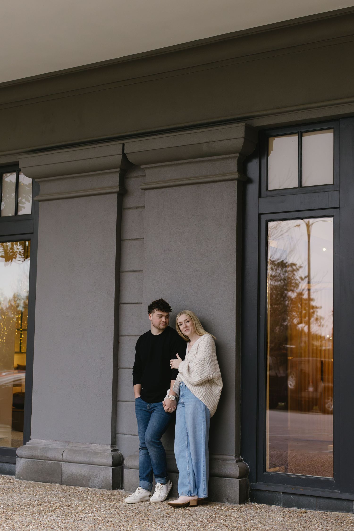 Couple in casual outfits posing against a modern building exterior with large windows.