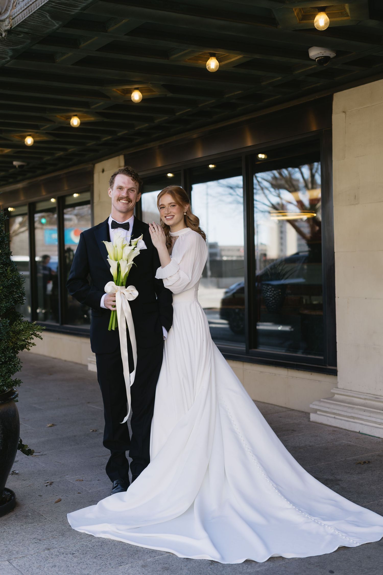 A bride in a flowing white gown and groom in a black tuxedo pose together under outdoor lighting.