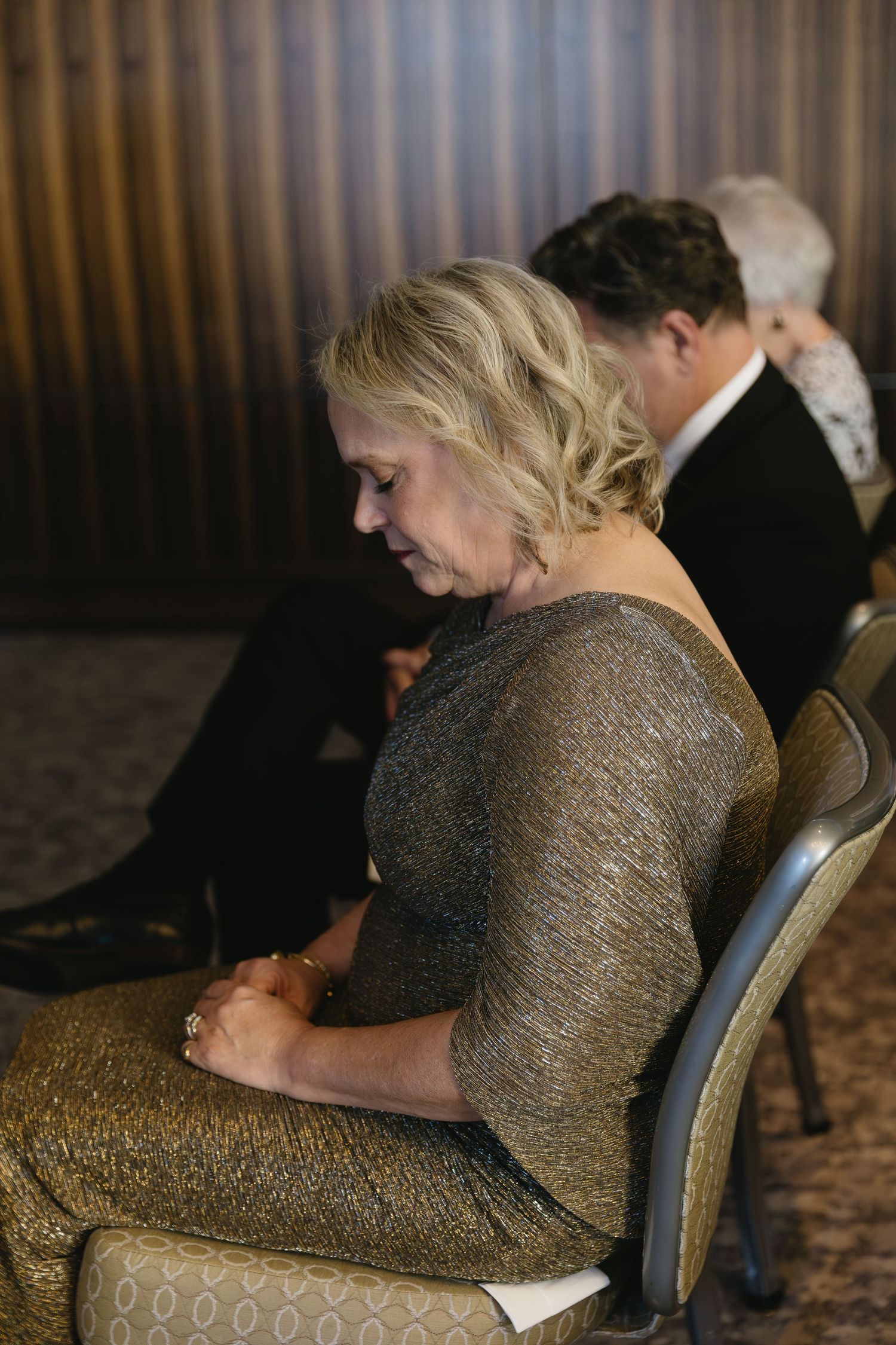 People seated in formal attire at an indoor event with warm lighting and textured walls.