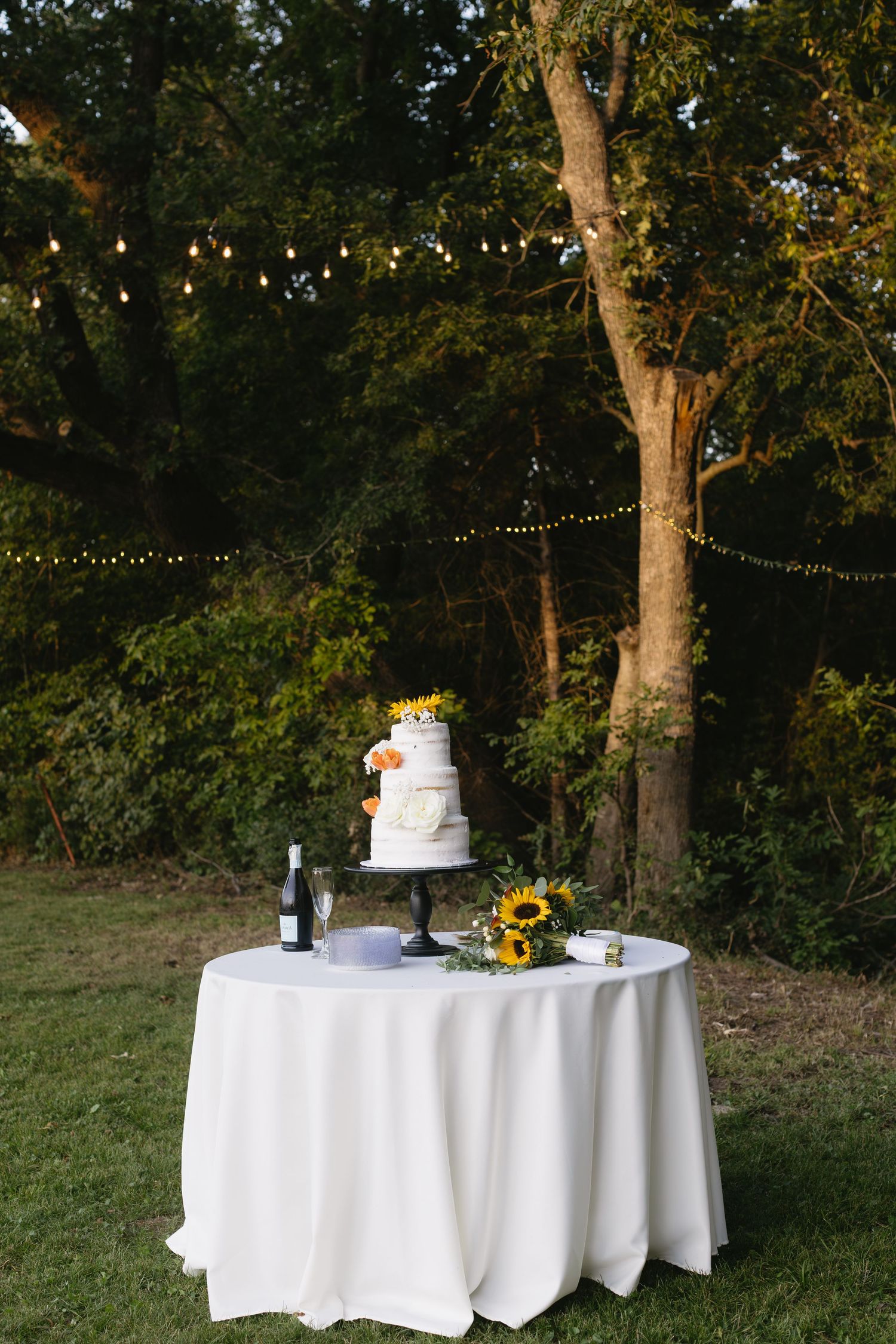 Round tables with white tablecloths and wedding cakes displayed in an outdoor garden setting at sunset.