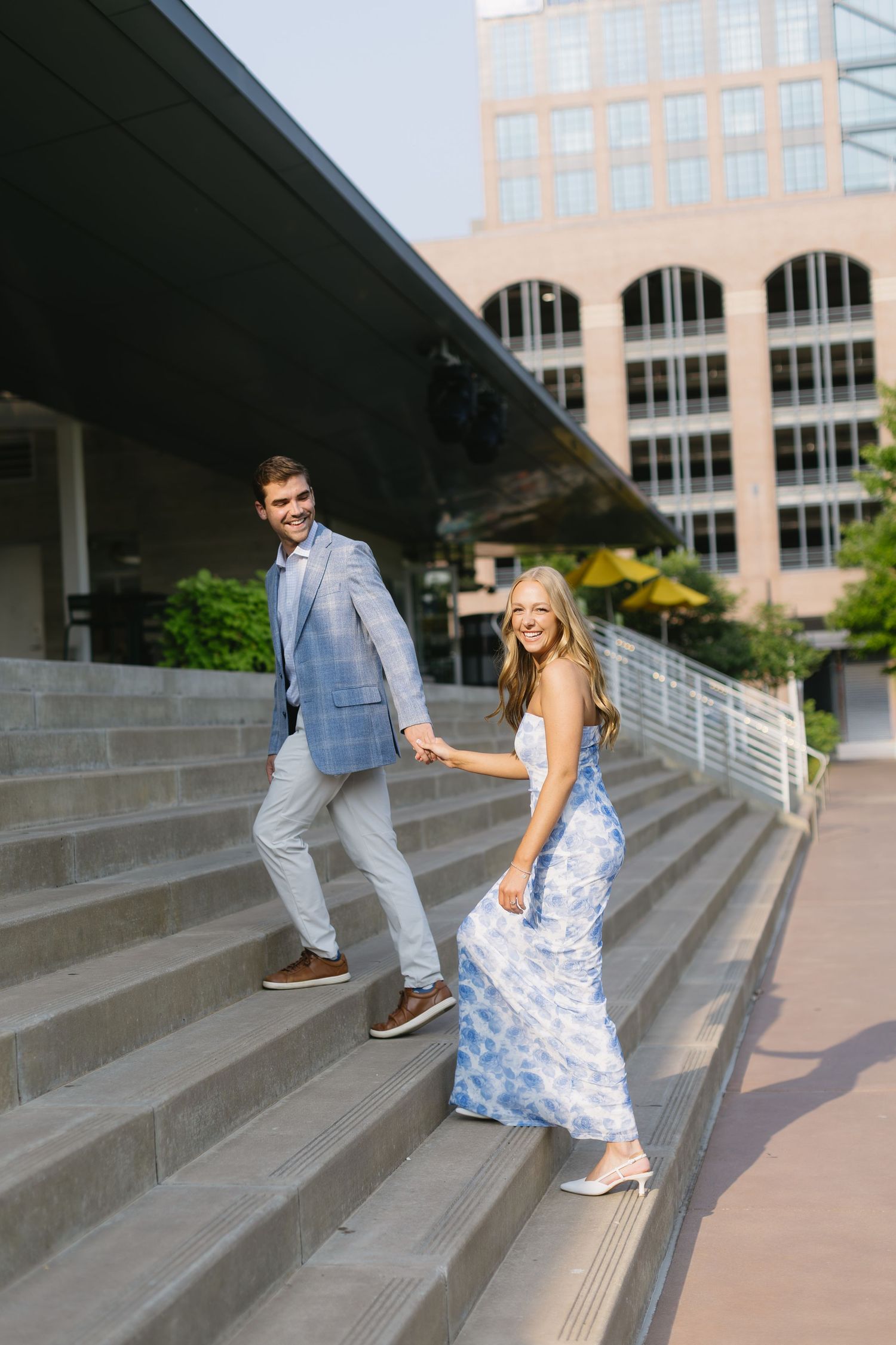 A couple walks up concrete steps wearing formal attire with a blue and white floral dress and light blue suit.