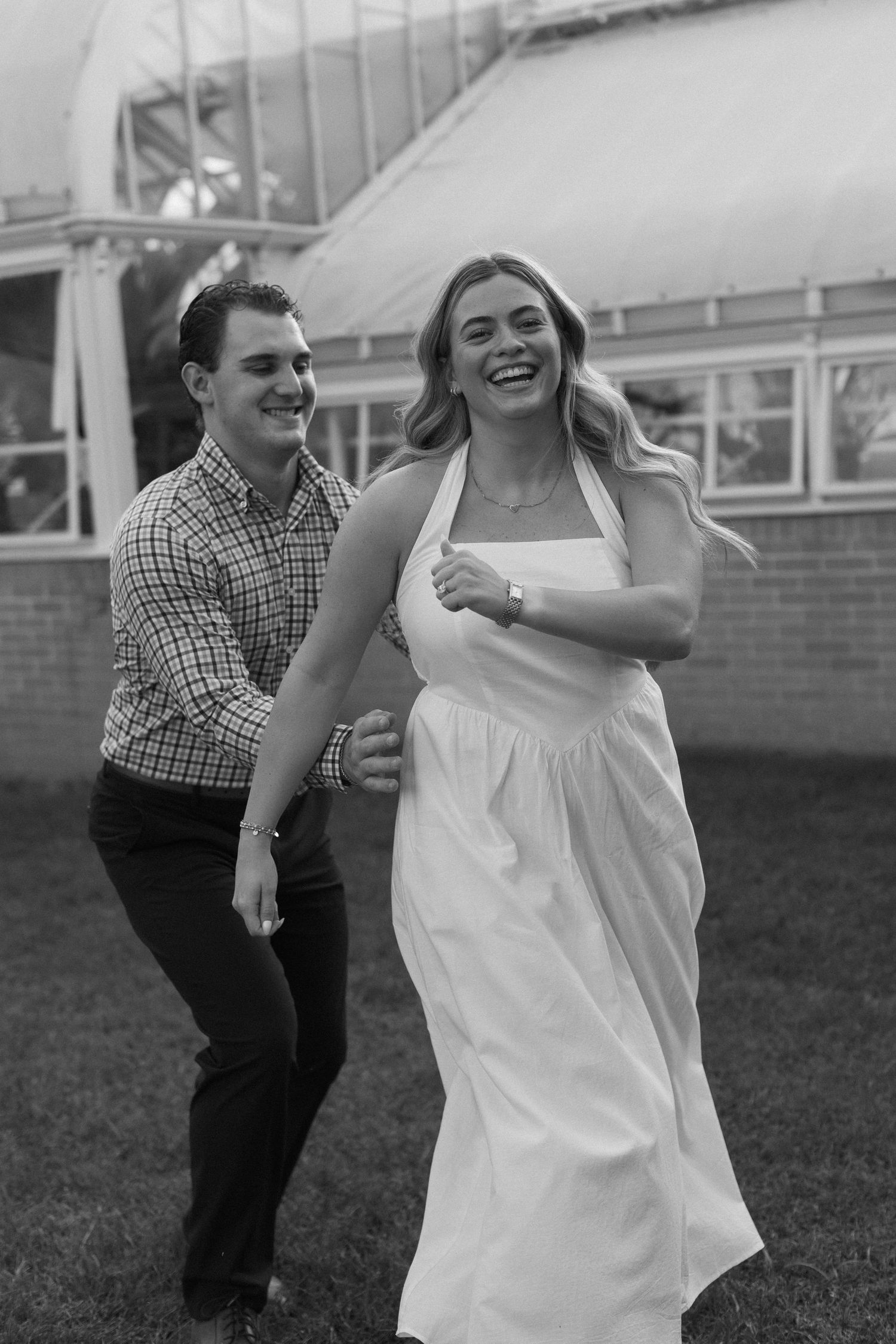 A joyful couple dances and laughs together on a grassy area near a brick building.
