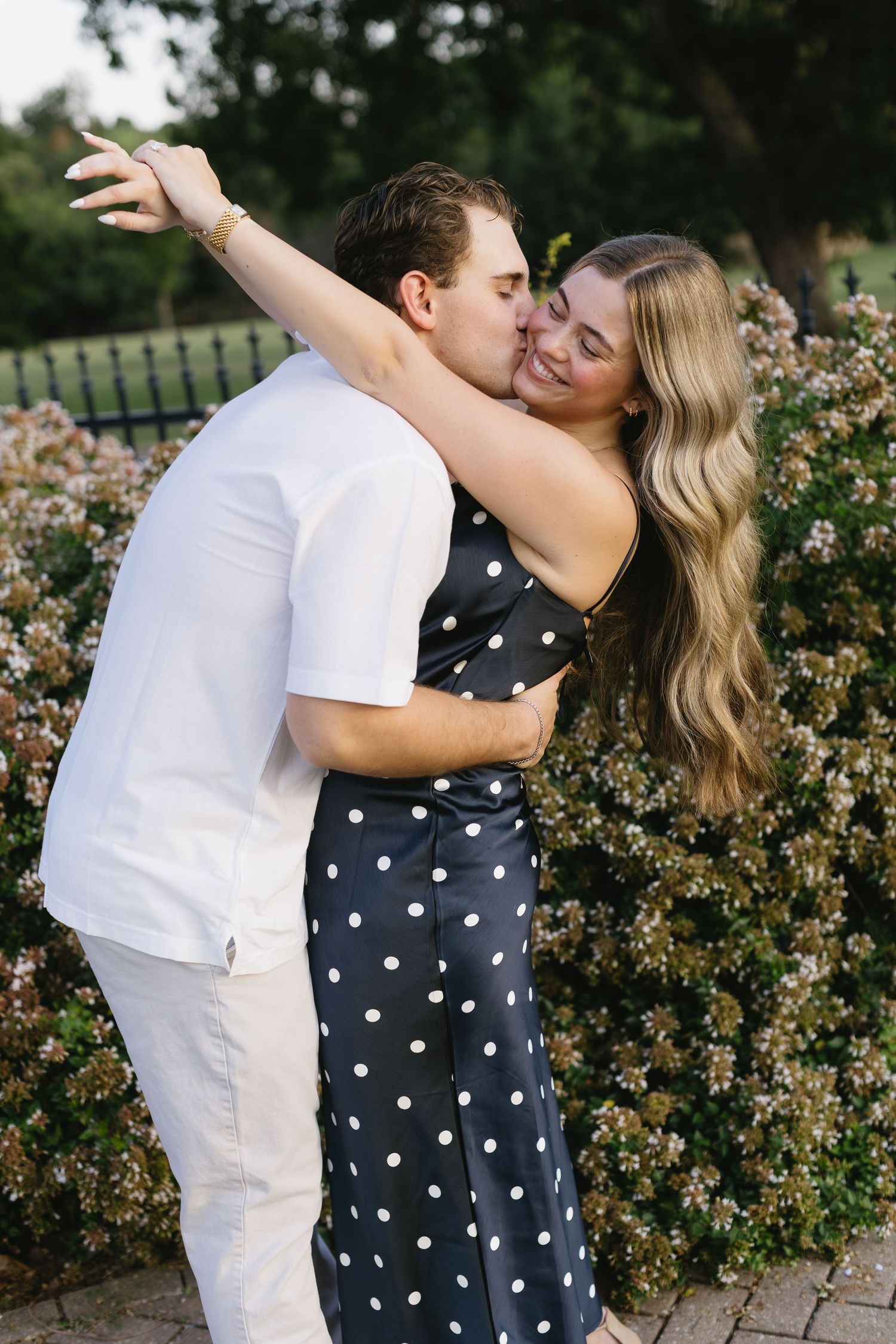 Couple in polka dot dress and white outfit embrace near blooming rose bushes in garden setting.