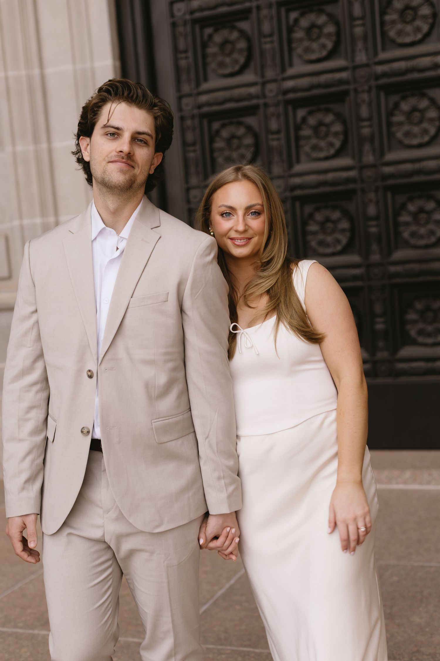 A couple in coordinated beige and white formal attire stand together against a decorative dark background.