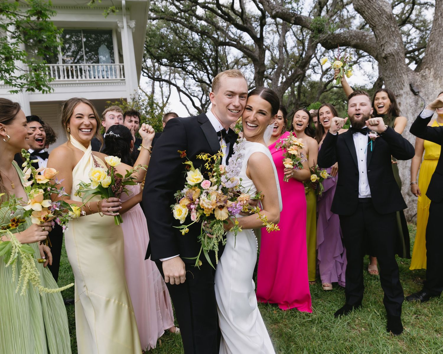 Wedding party poses outdoors with bridesmaids in colorful dresses holding bouquets.