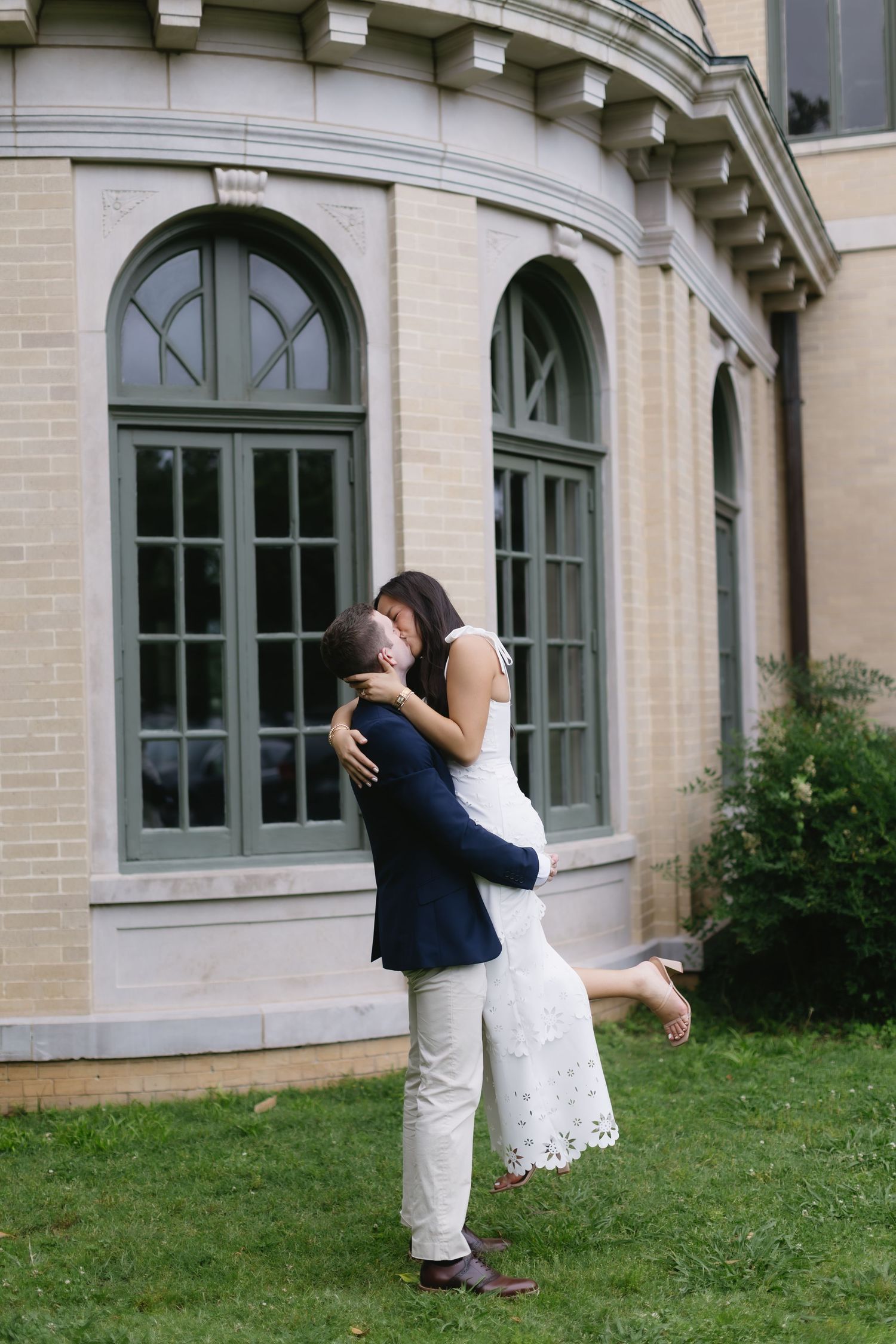 Playful couple dips and embraces in front of elegant stone building with arched windows.