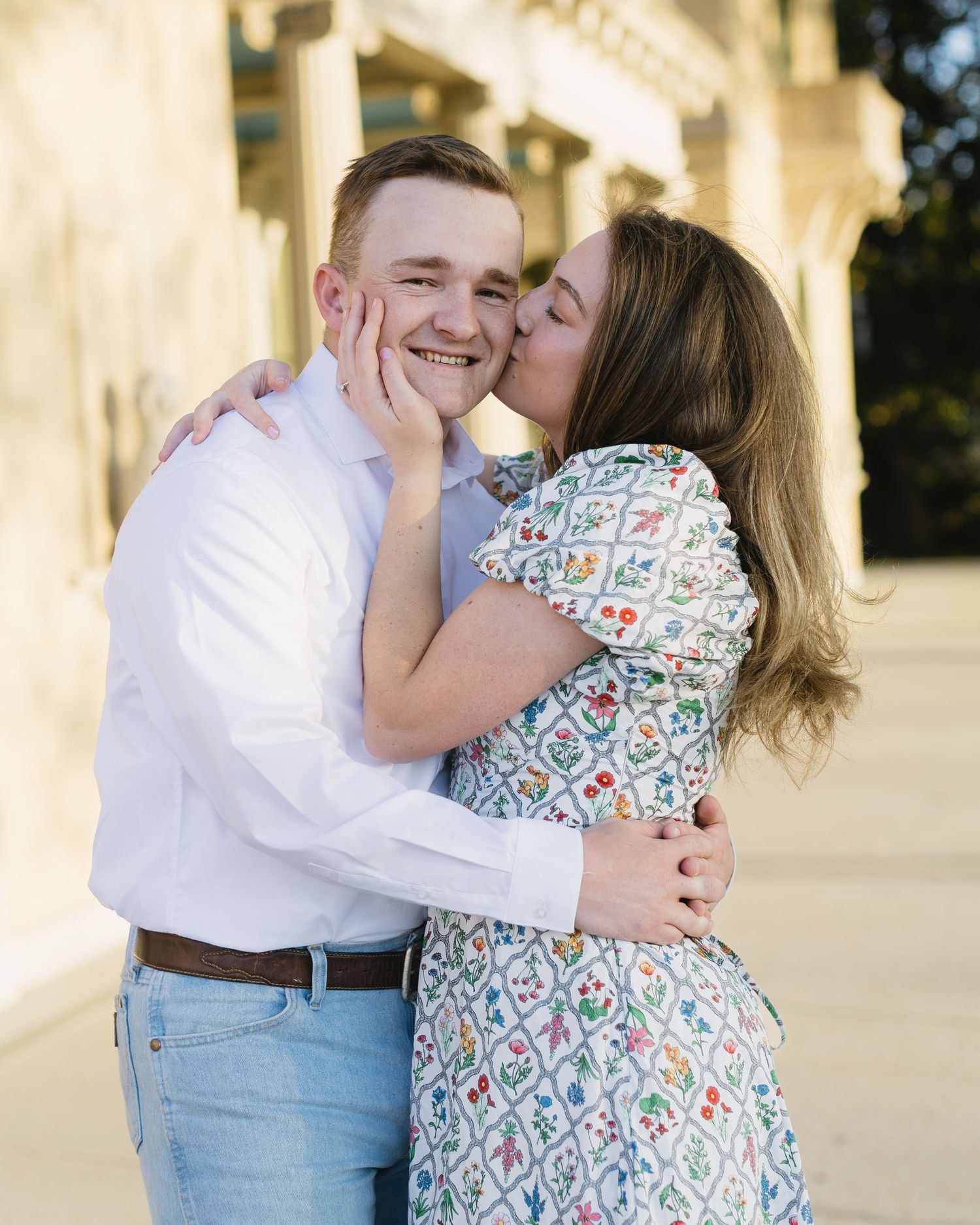 A romantic couple embraces and laughs together near white pillars at sunset.