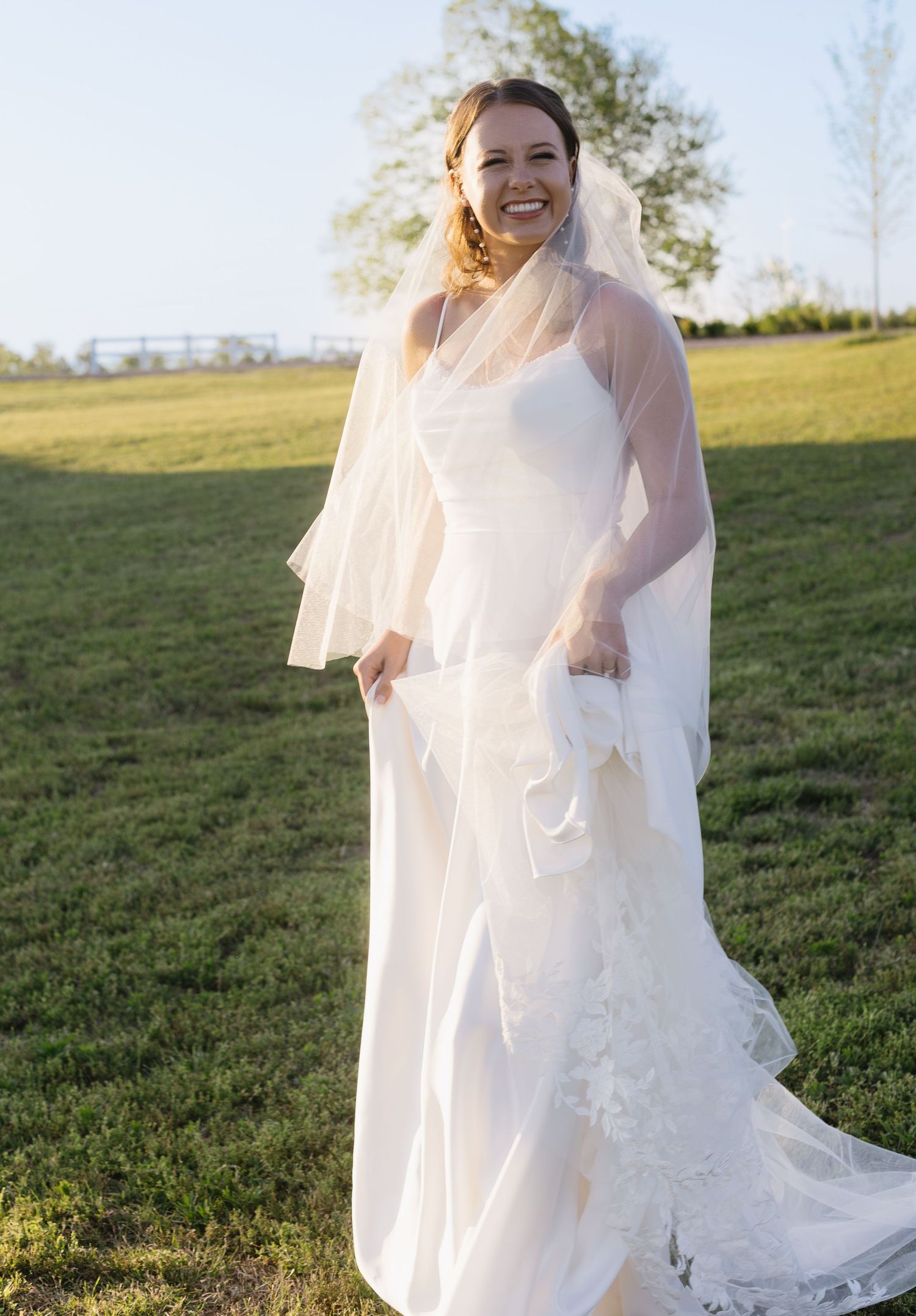 A bride dances and twirls in her wedding dress on a grassy hillside during golden hour.