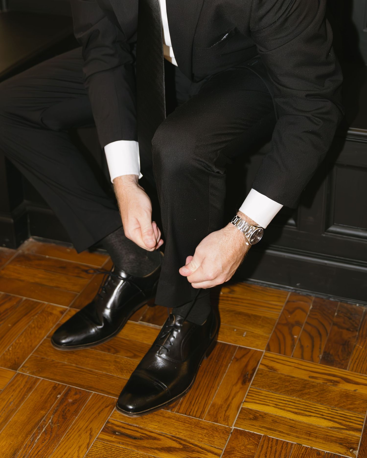 Close-up of formal black dress shoes and suit pants on parquet flooring.