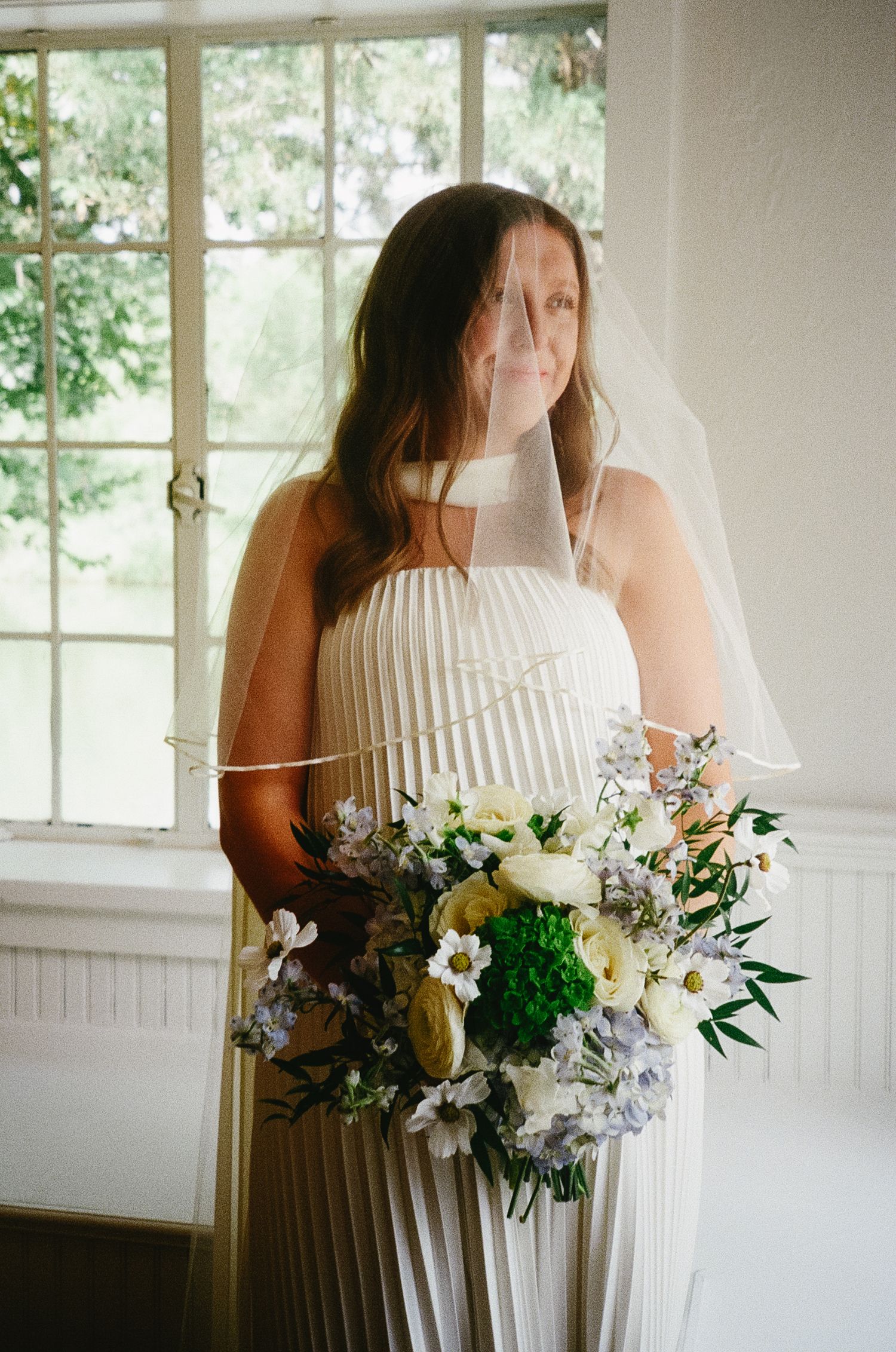 A bride in a floral lace dress holds a beautiful white and blue bouquet of flowers in a sun-lit room with windows.