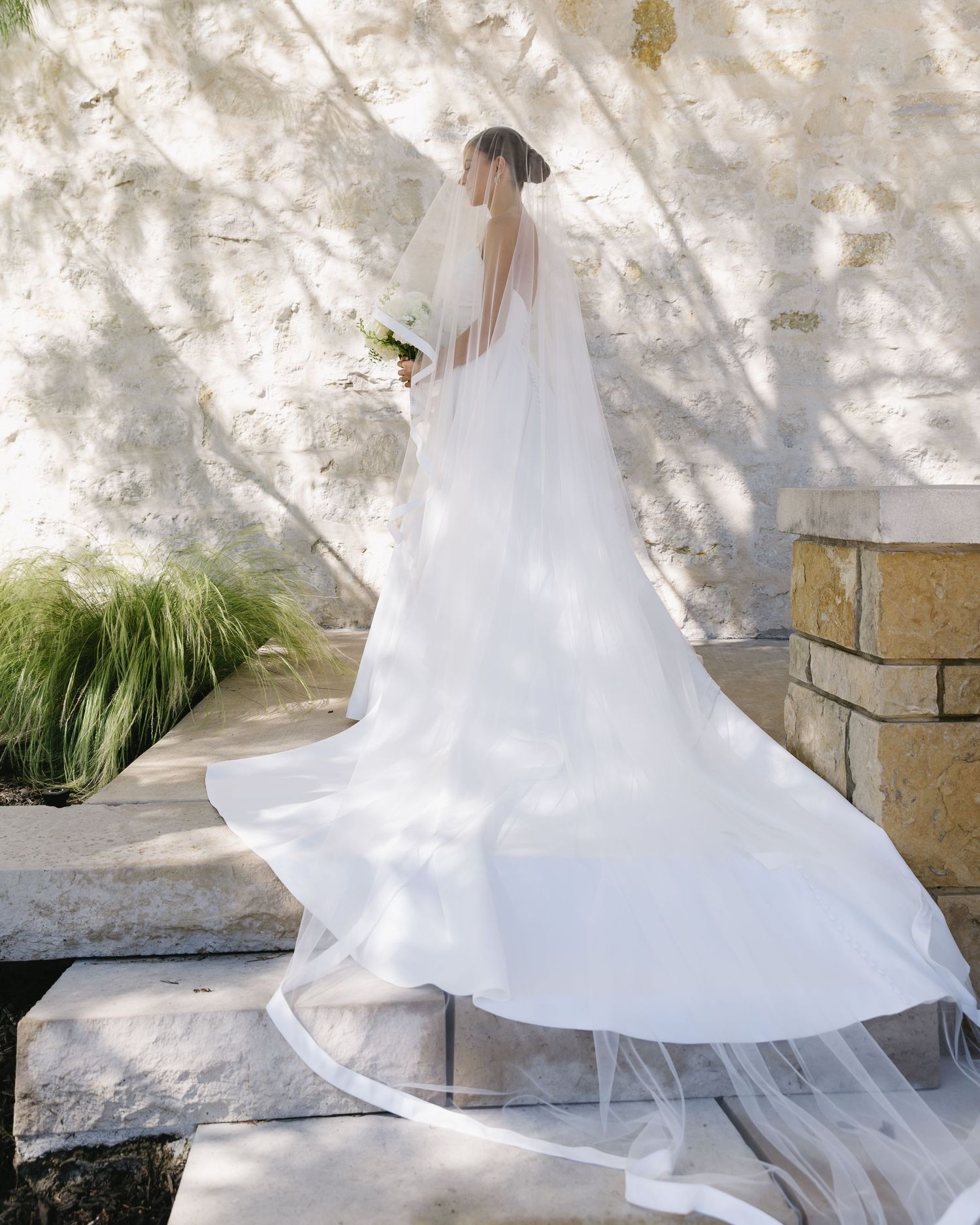 Flowing white wedding dress train moving in the breeze against stone wall backdrop.