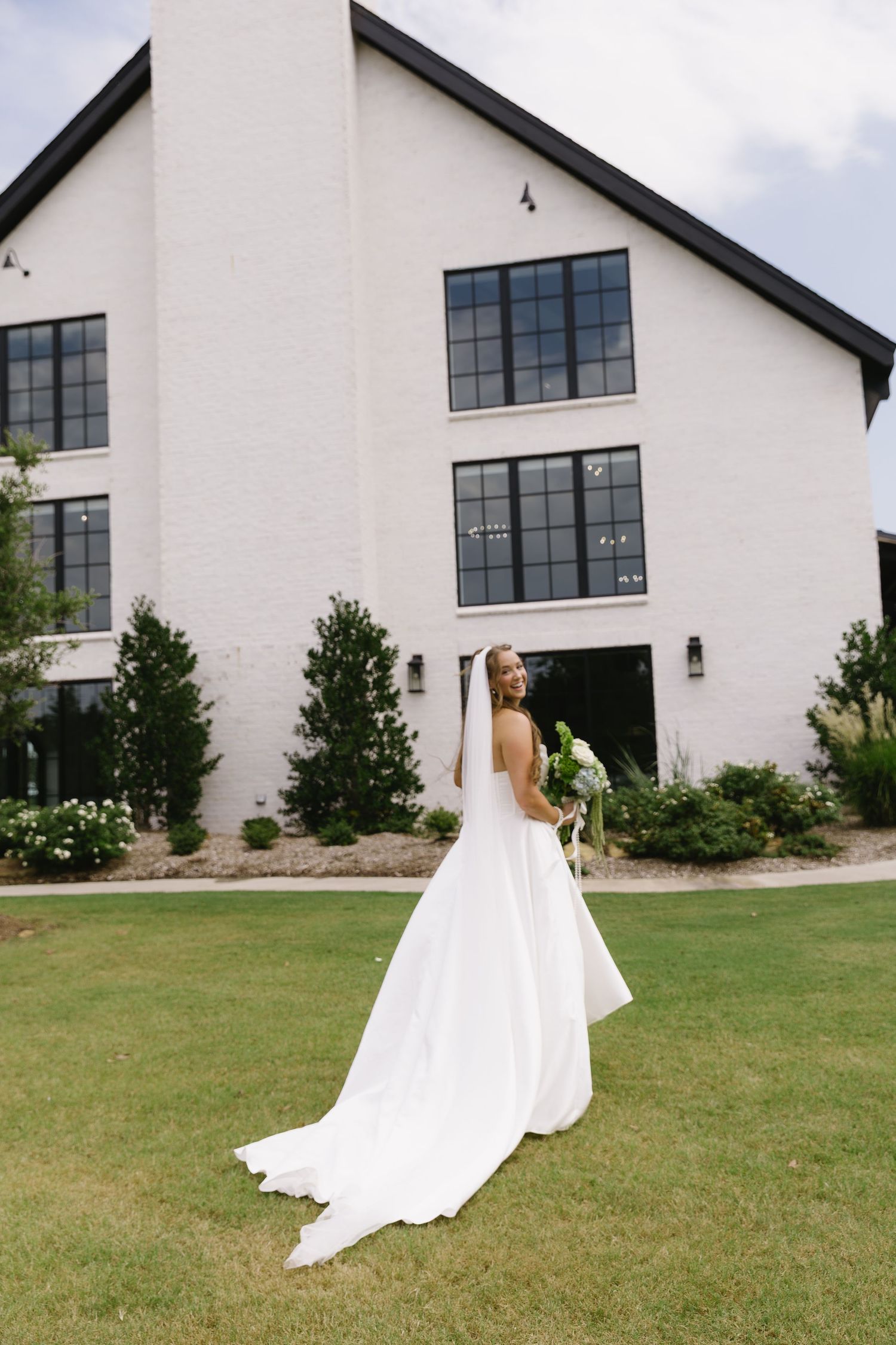 A bride in a flowing white dress poses in front of a modern white building with large windows.