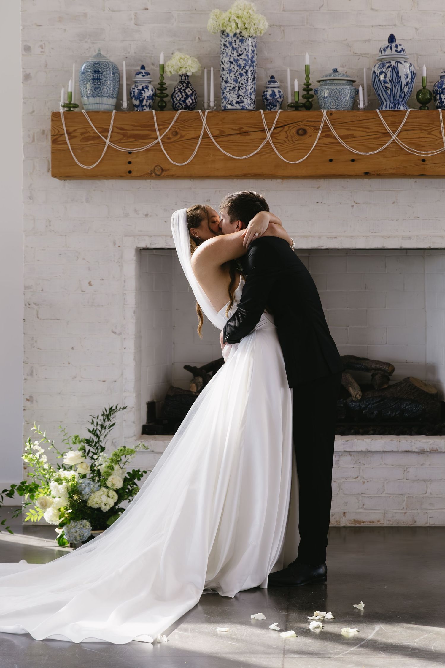 Romantic wedding couple embraces in front of white brick fireplace with floral decor.