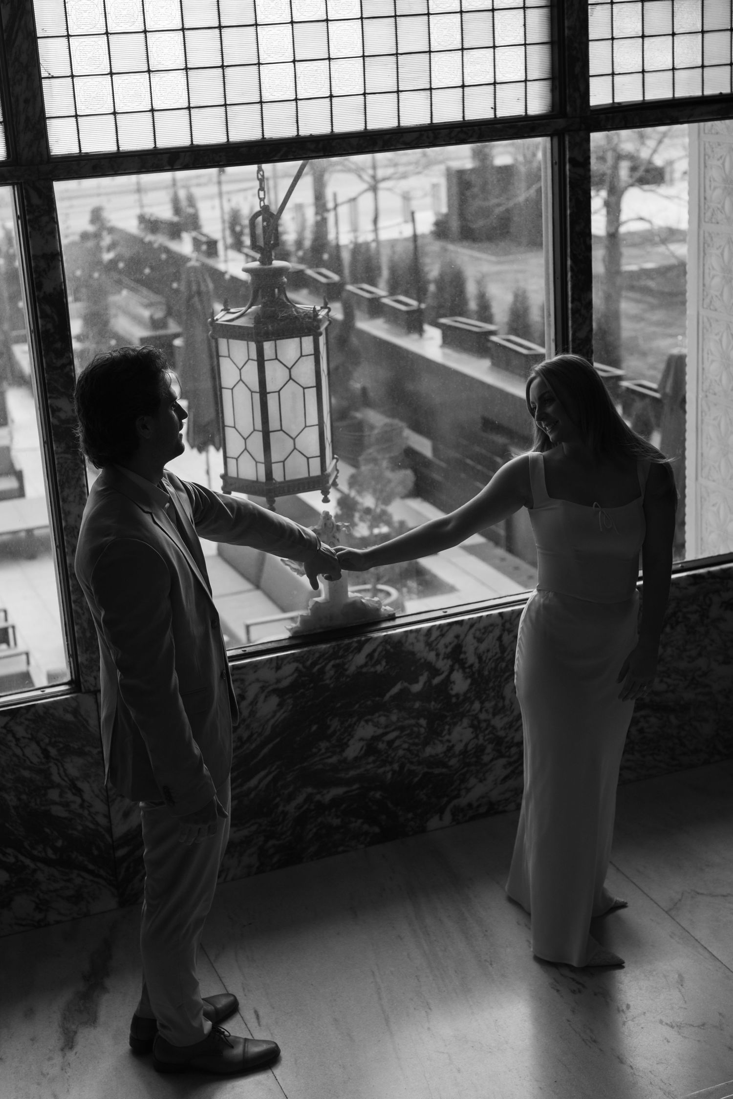 A black and white photo of a couple in formal wear standing by a large window with geometric panes.