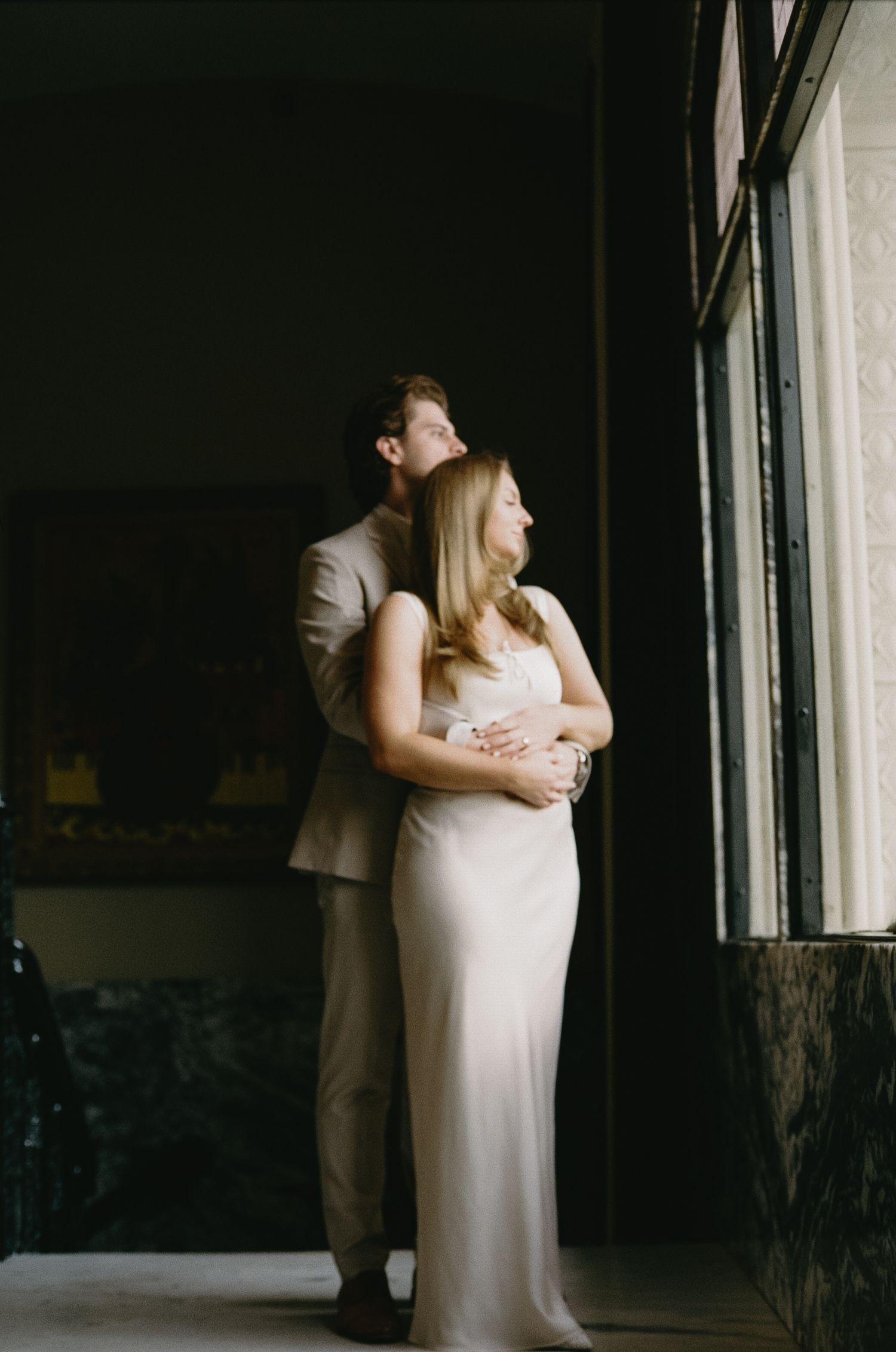 A couple in white and beige embraces in dramatic lighting near a window.