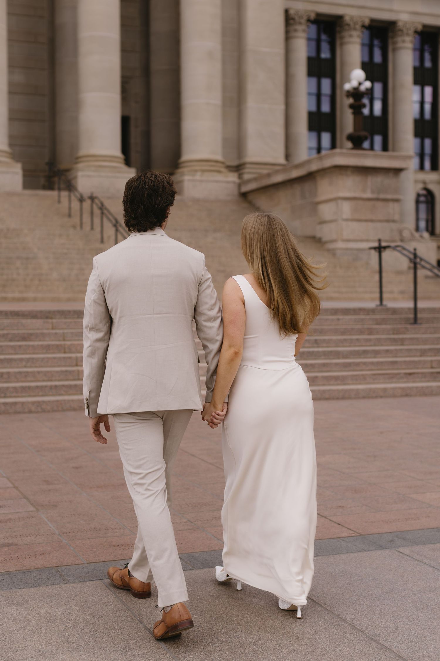 A couple in matching light-colored formal attire walks up stone steps holding hands at a grand building entrance.