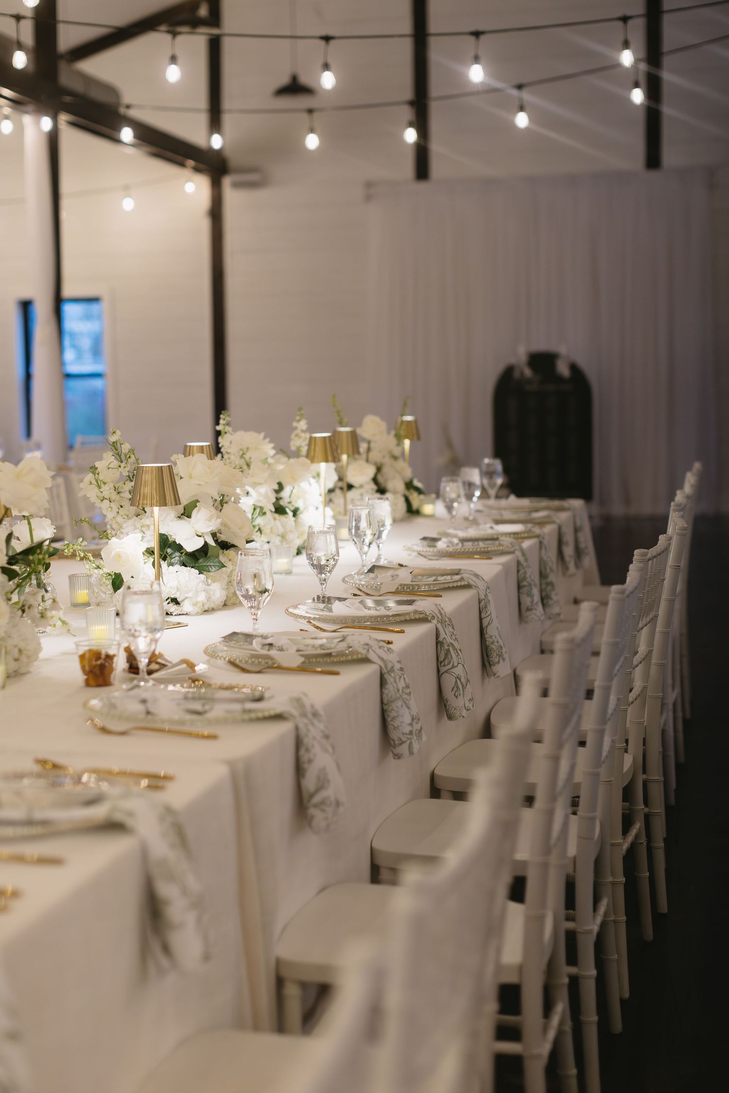 Elegant wedding reception tables set with white linens and chairs in a modern venue with string lights.