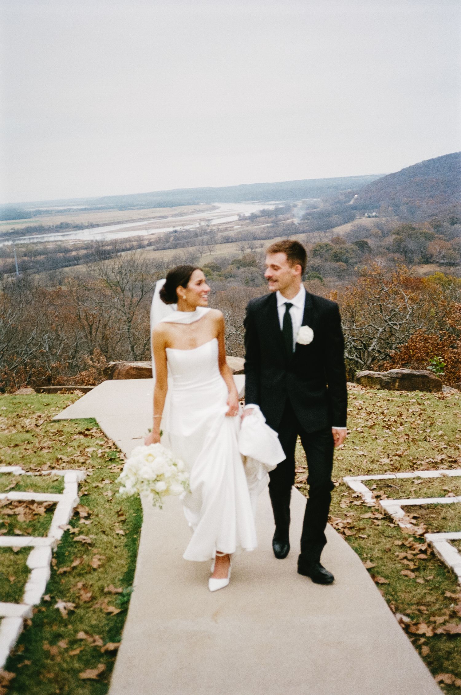 Couple in wedding attire sitting on steps with scenic mountain view in background.