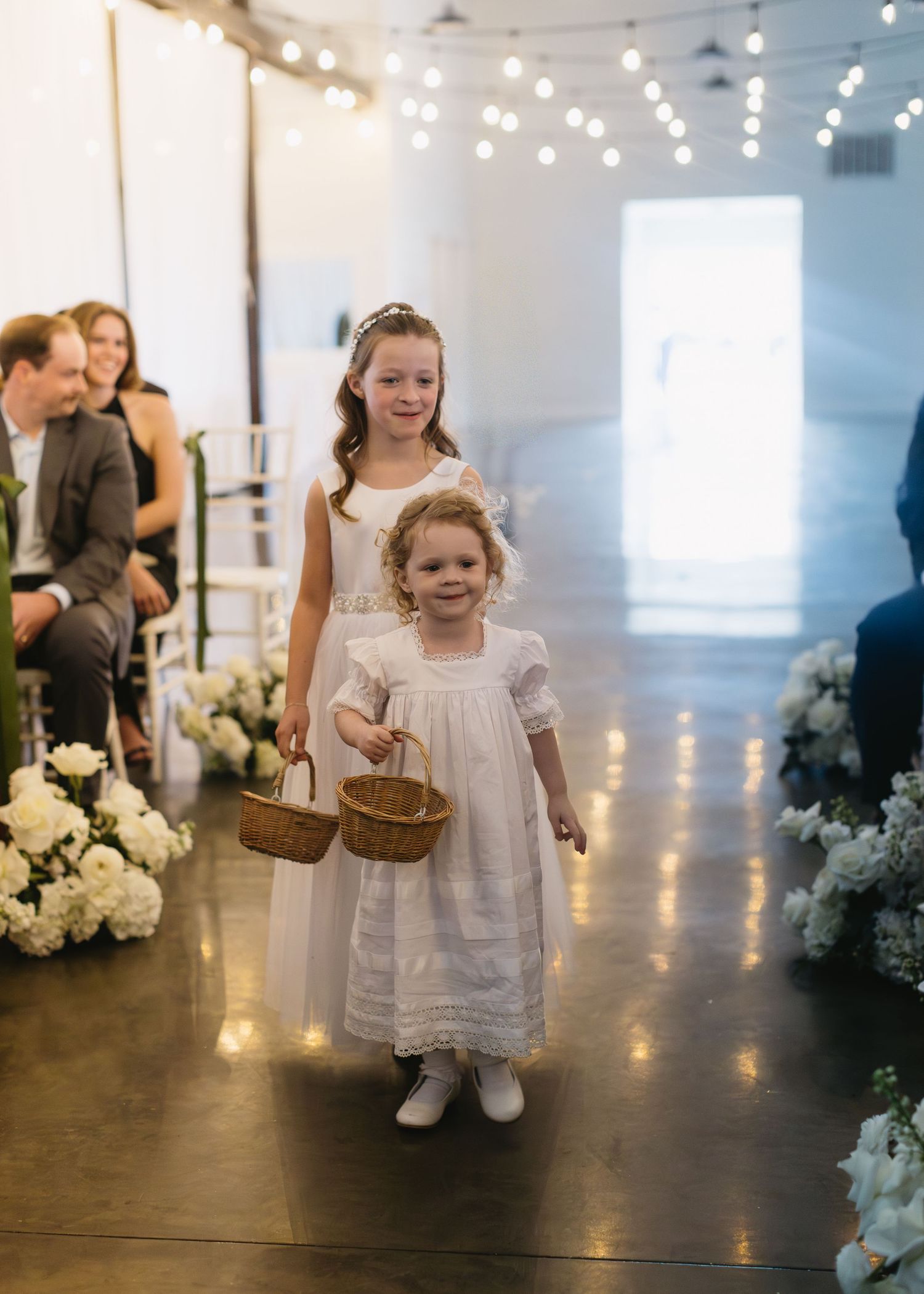 Young flower girls in white dresses walk down the aisle carrying baskets at a wedding ceremony.