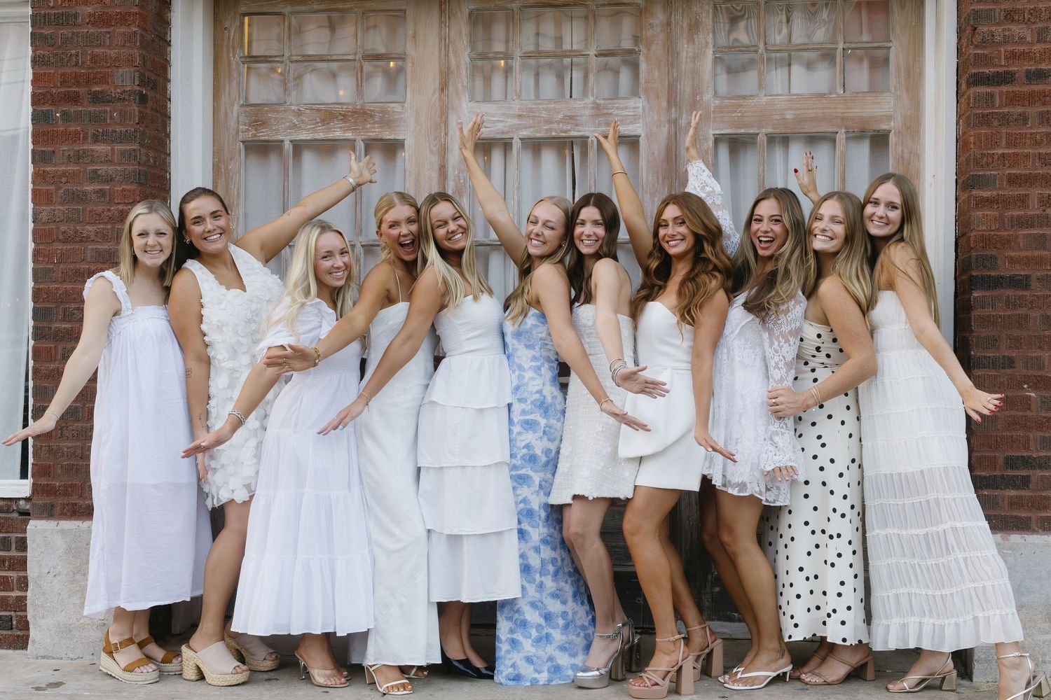 Group of friends in white and floral dresses posing together against a brick wall with a vintage door.