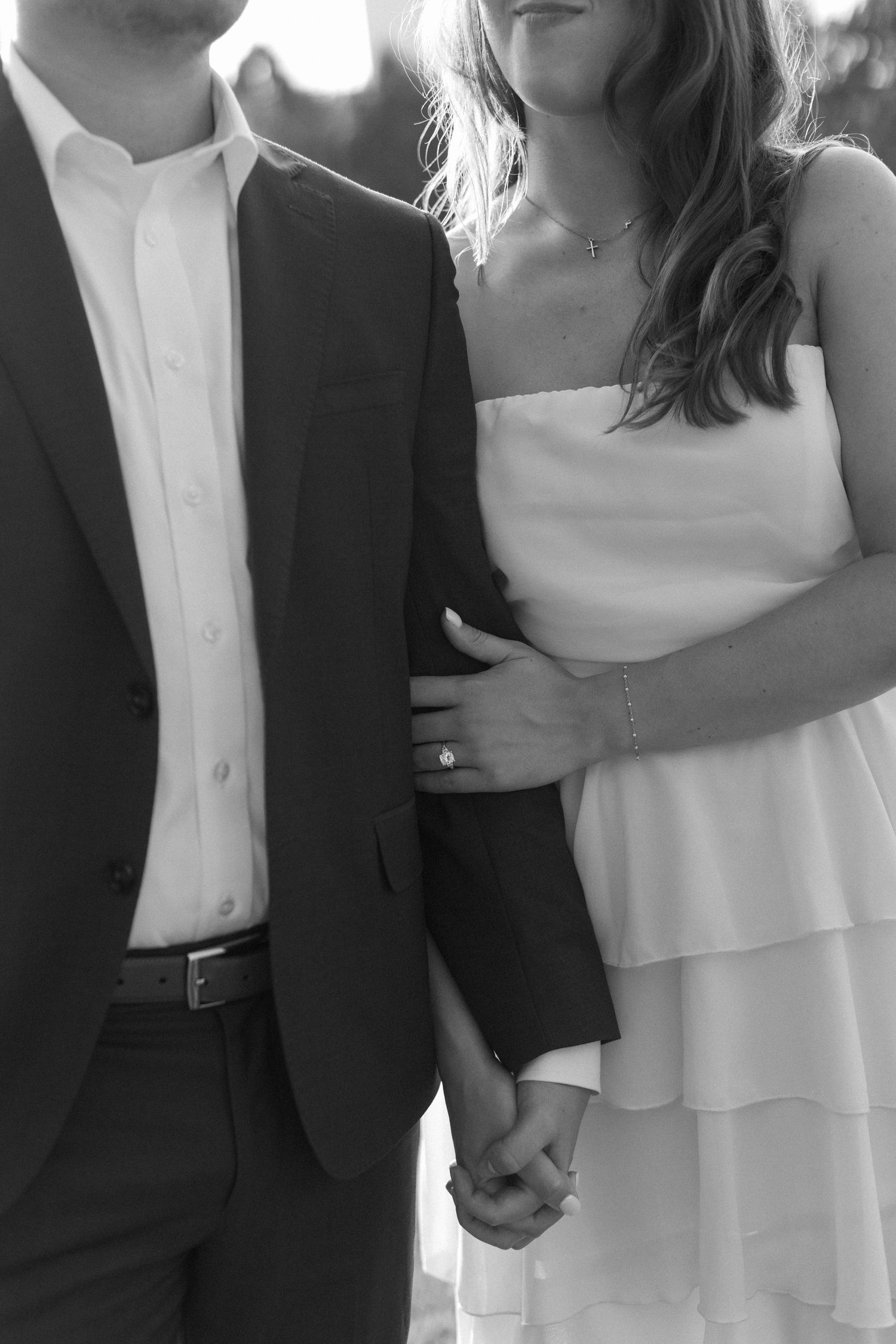 A couple in formal attire holds hands, with her wearing a tiered white dress and him in a black suit.
