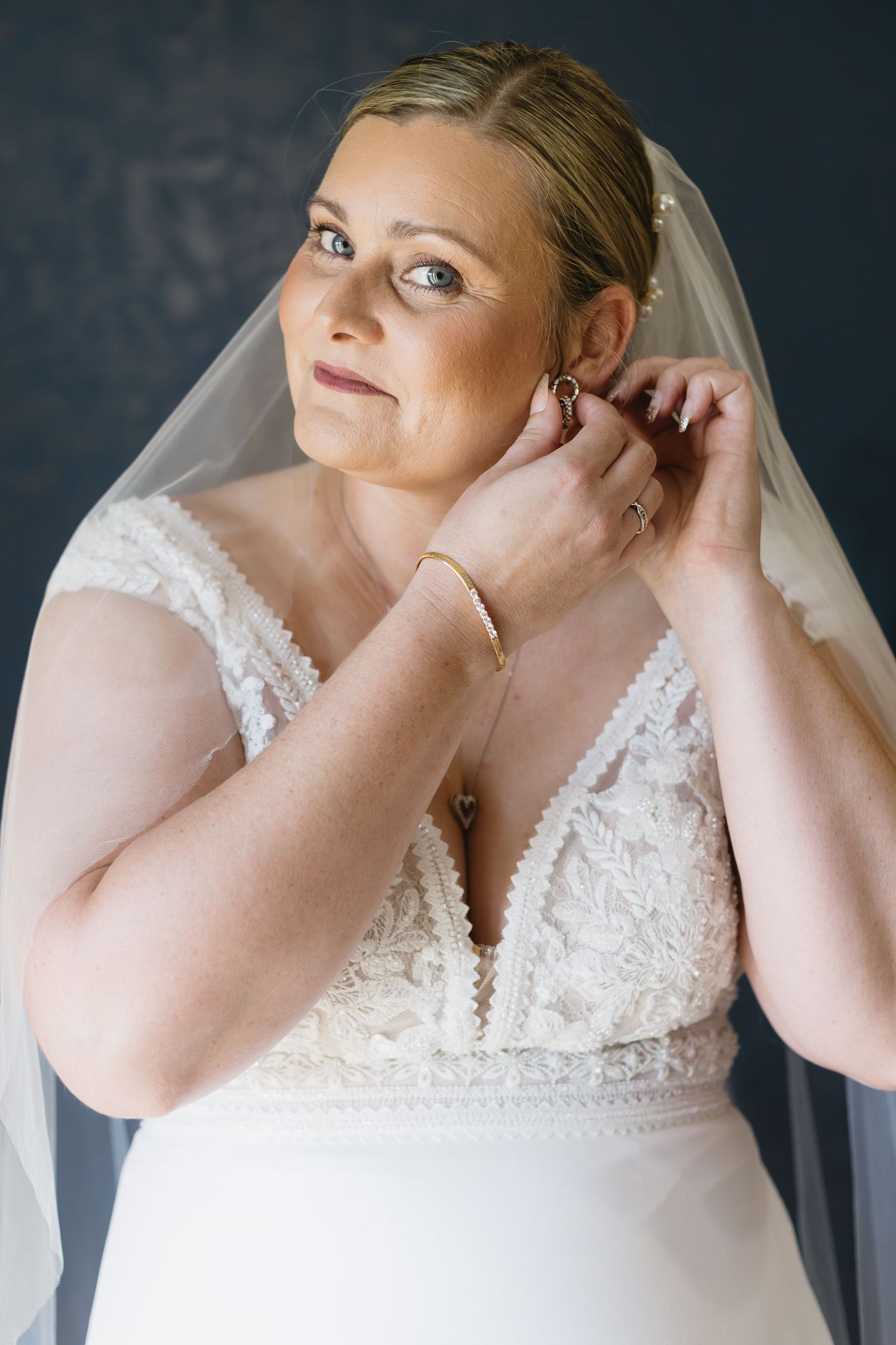 Bride wearing a delicate lace wedding dress and veil puts on earrings while getting ready against a dark backdrop.