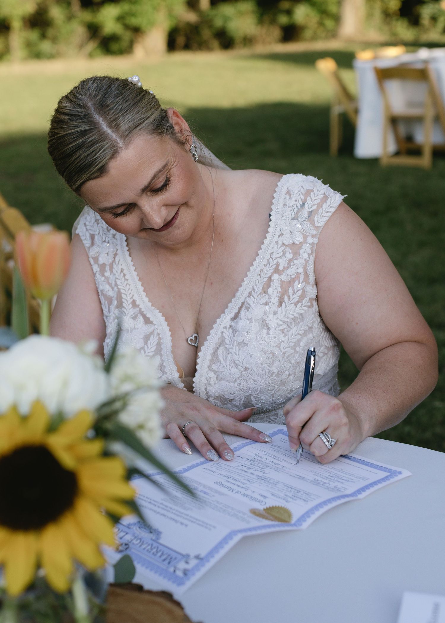 A bride in a lace dress signs marriage documents at an outdoor table with sunflower decorations.