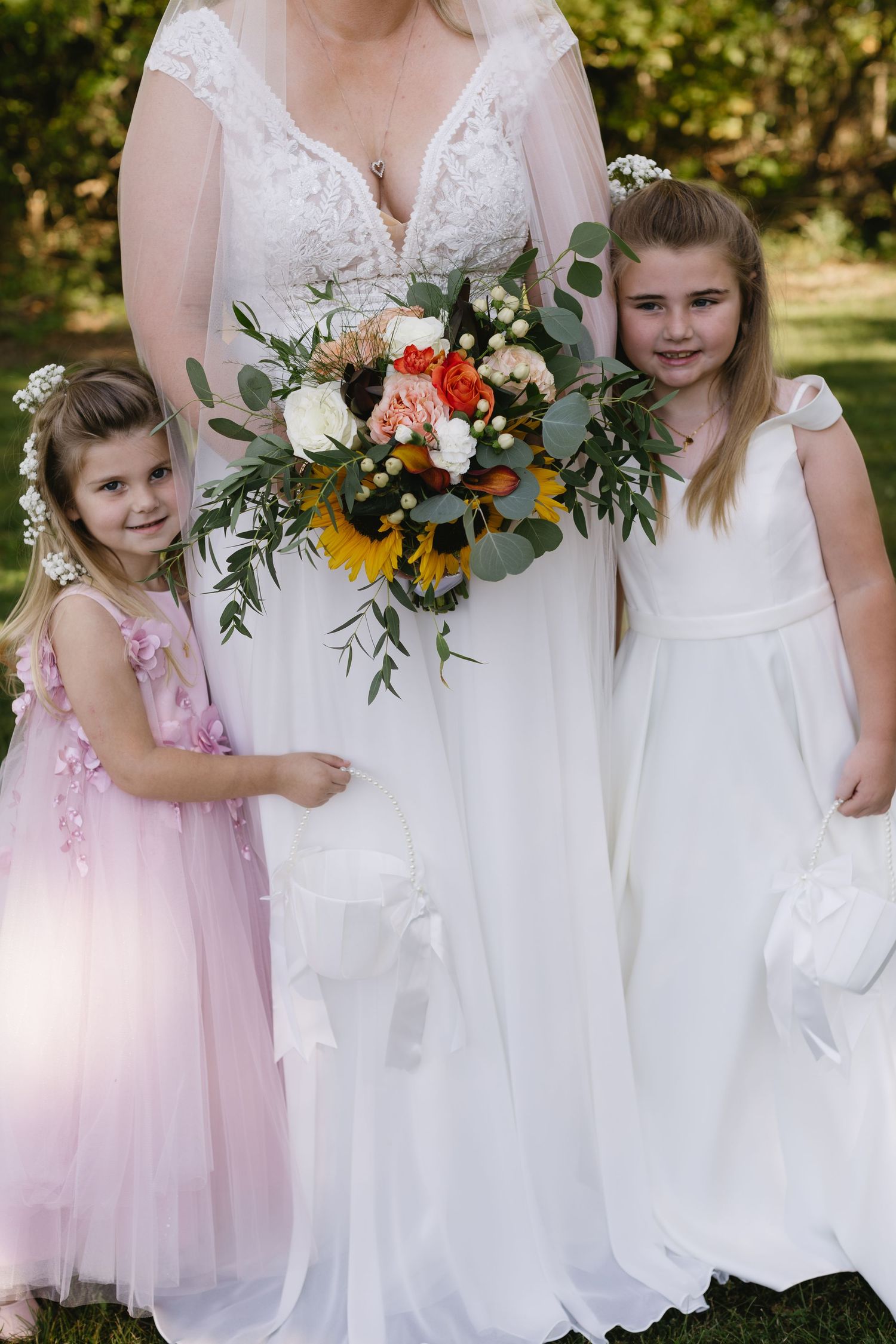 A bride in a white lace dress holds a colorful bouquet with sunflowers and roses next to someone in a pink dress.