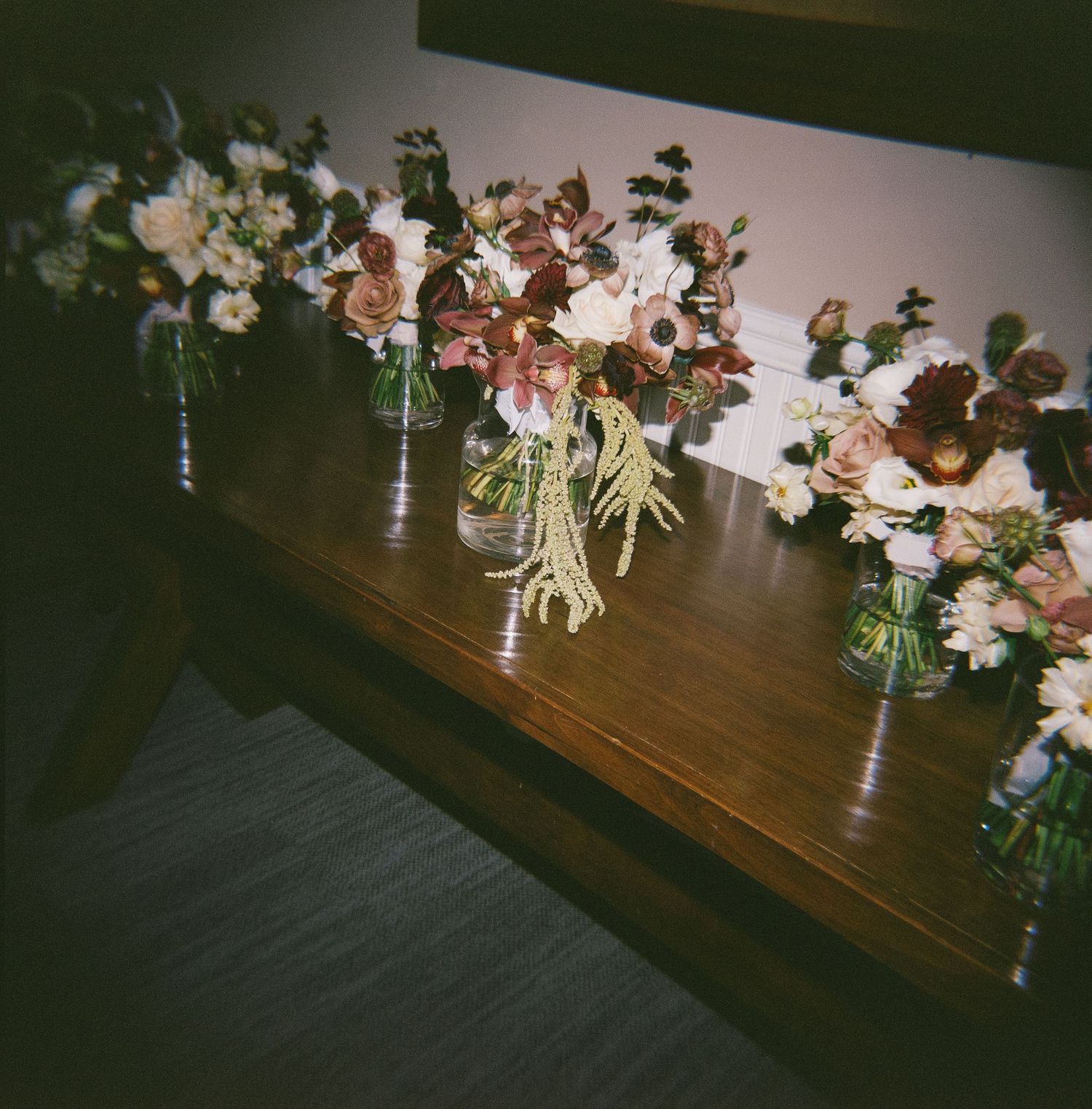 Floral arrangements in glass mason jars line up on a dark wooden table surface creating an elegant rustic display.