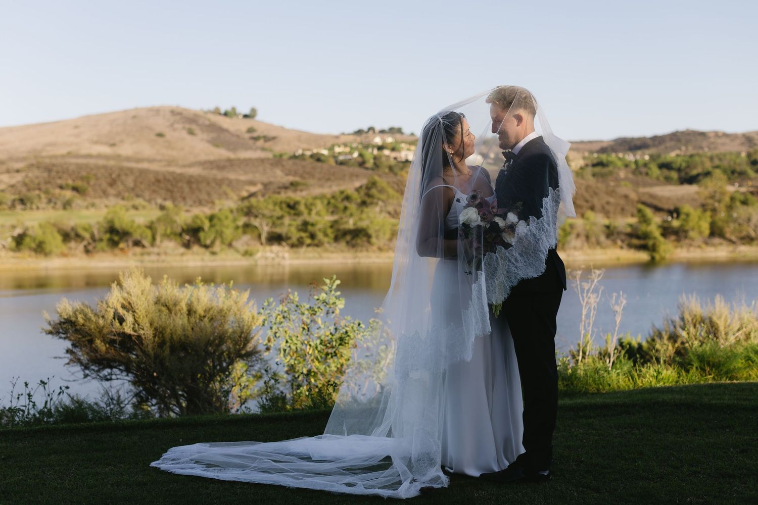 Wedding couple embracing in front of a scenic lake and rolling hills landscape.