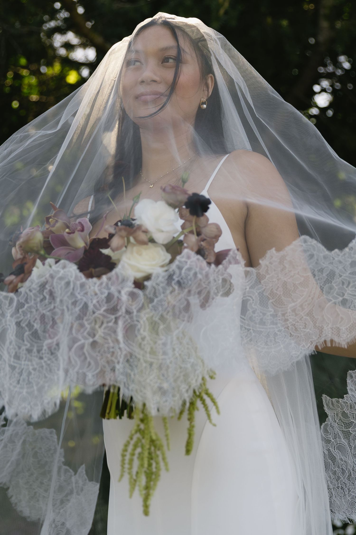 Bride wearing a delicate lace-trimmed veil holding a romantic burgundy and white floral bouquet.