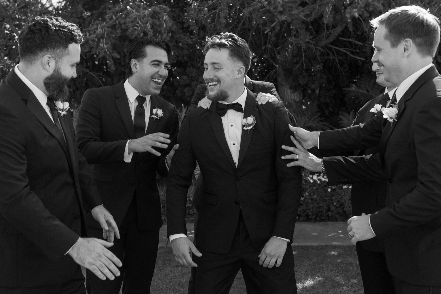 Wedding party members in formal attire share enthusiastic greetings outdoors.