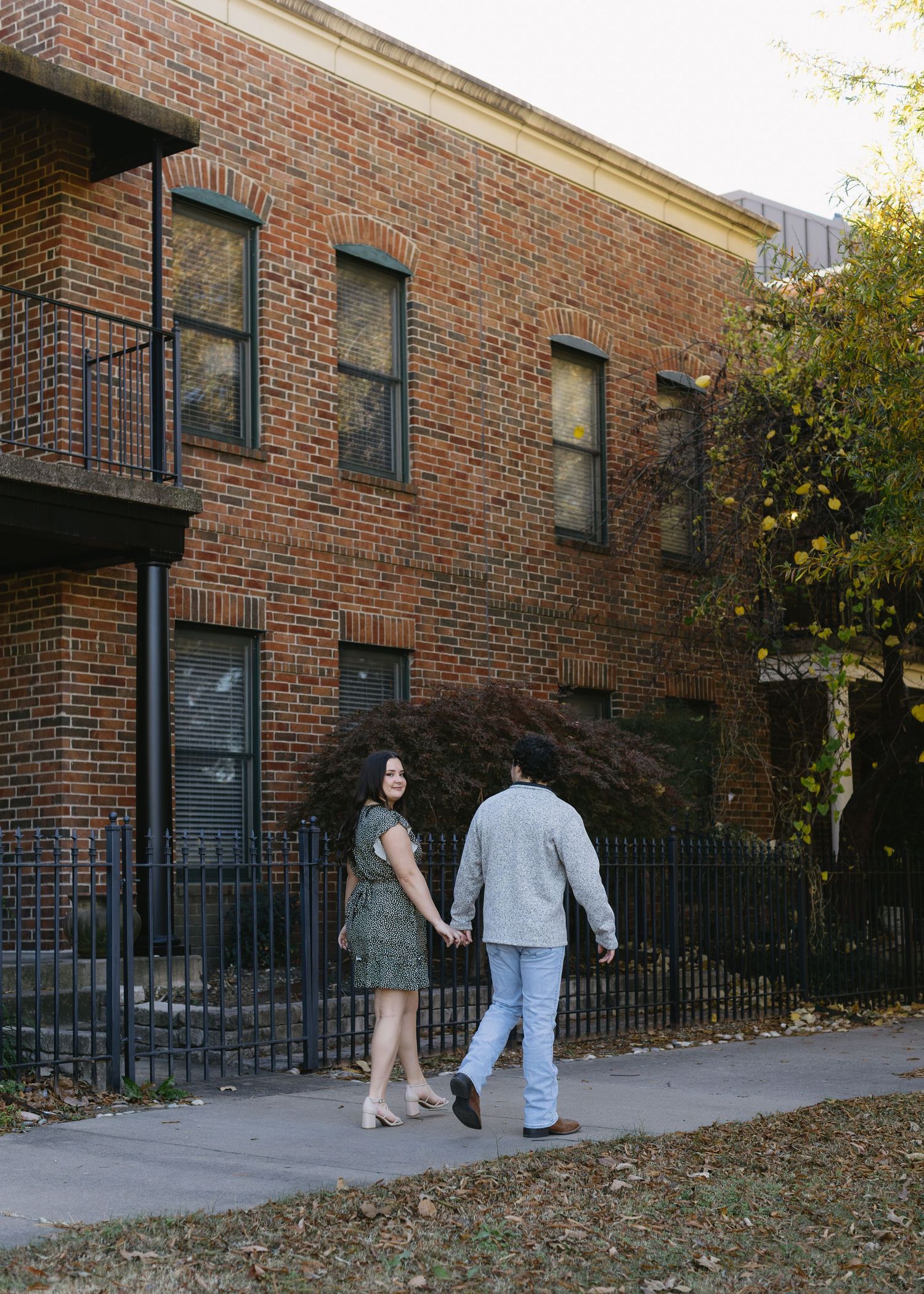 A couple strolls together on a walkway beside a red brick apartment building with black iron railings.