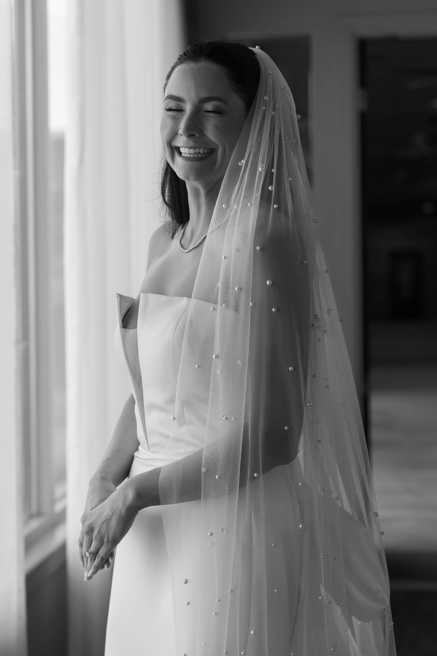A bride wearing a delicate veil with pearl accents poses in natural window light.