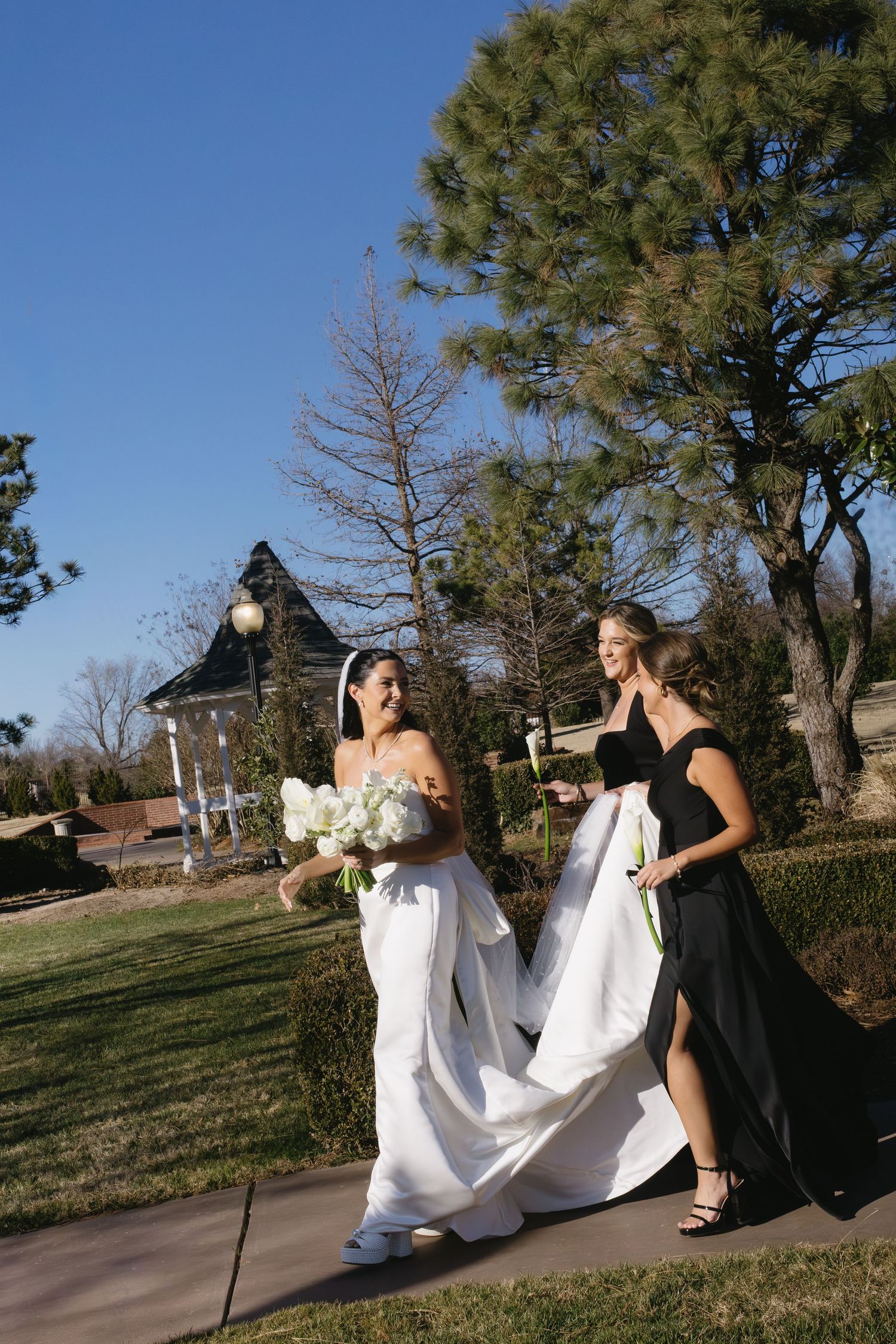 Bridesmaids in black dresses help adjust a white wedding gown train on a sunny outdoor path.