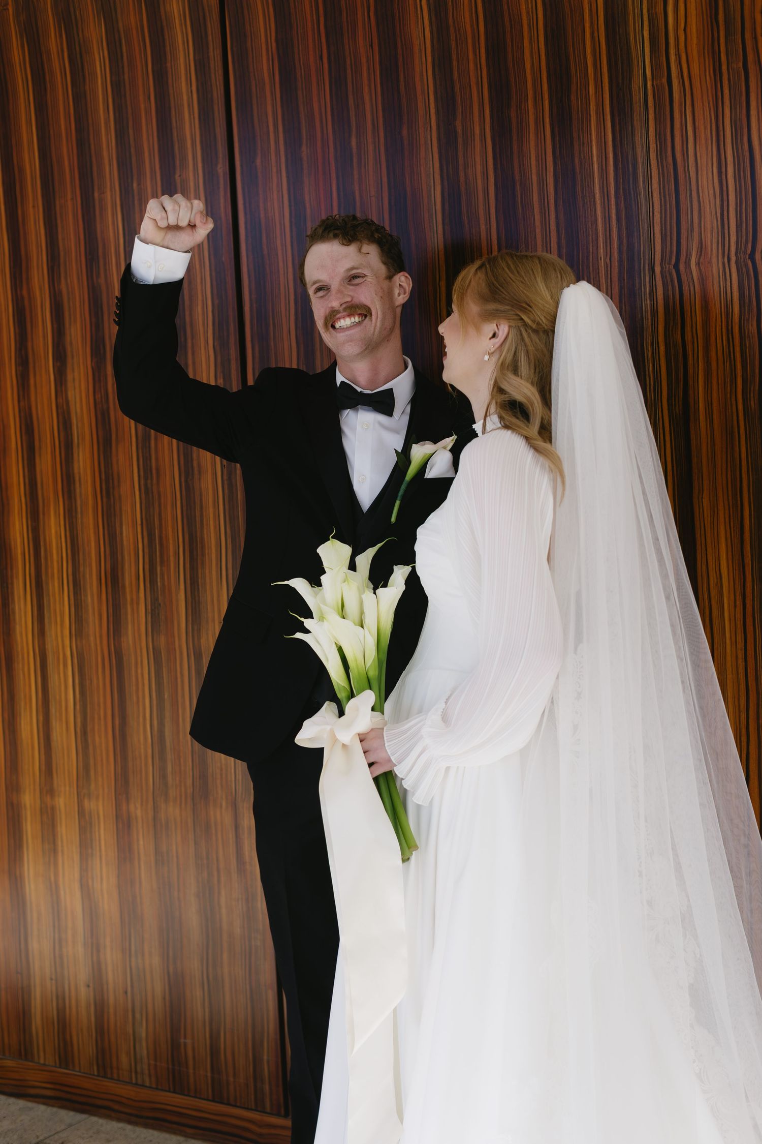 Newlyweds share joyful moment with white calla lilies against wooden backdrop.