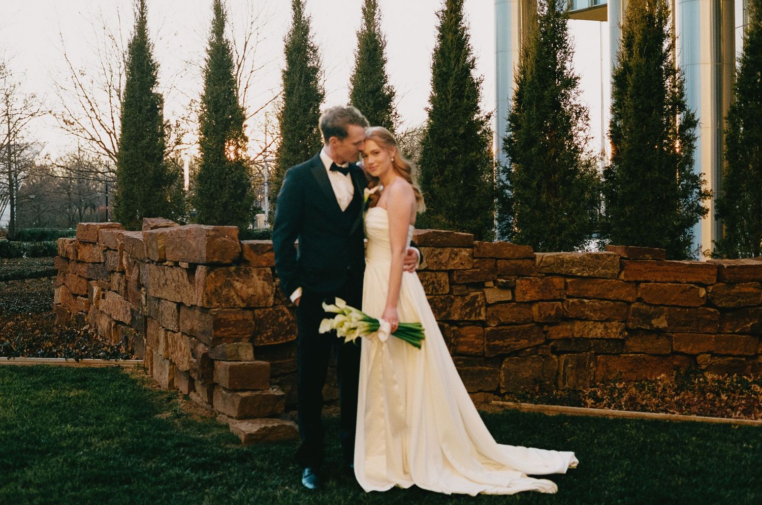 Wedding party poses along a stone wall with evergreen trees in the background.
