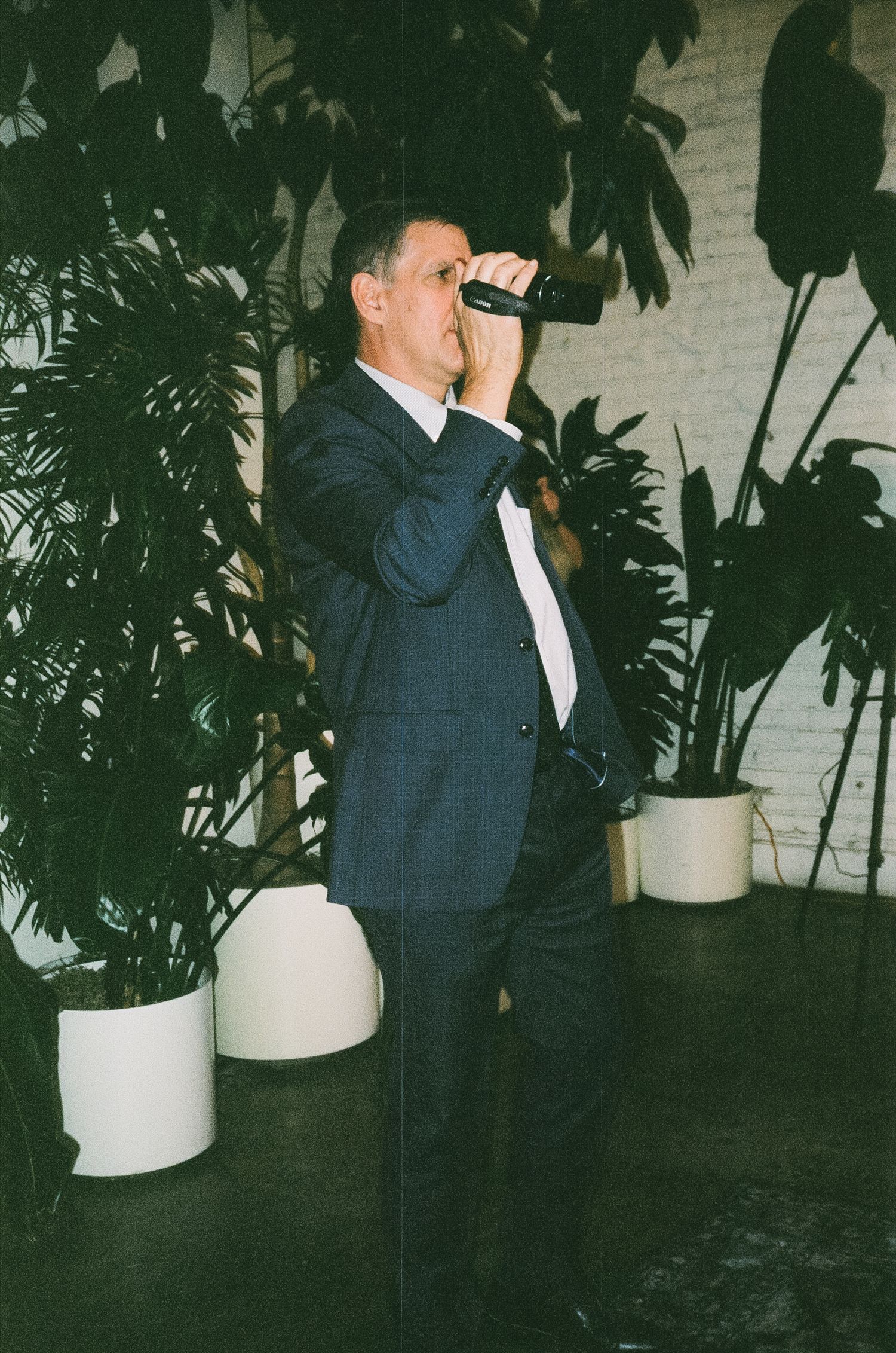 A guest in a navy suit takes a drink at a wedding reception with tropical plants in the background.