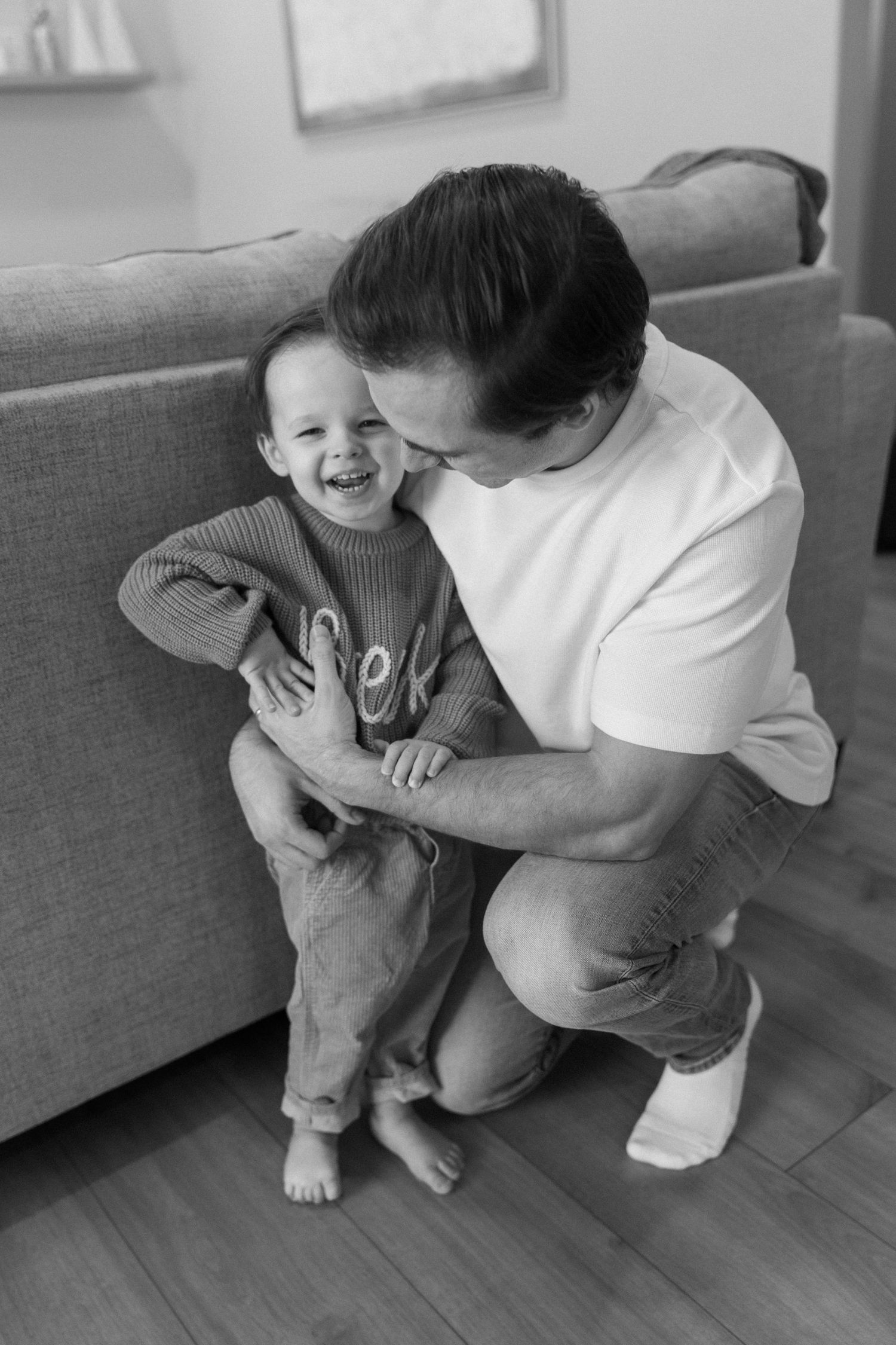 A tender moment between parent and child sharing laughter while sitting on a hardwood floor by a gray couch.