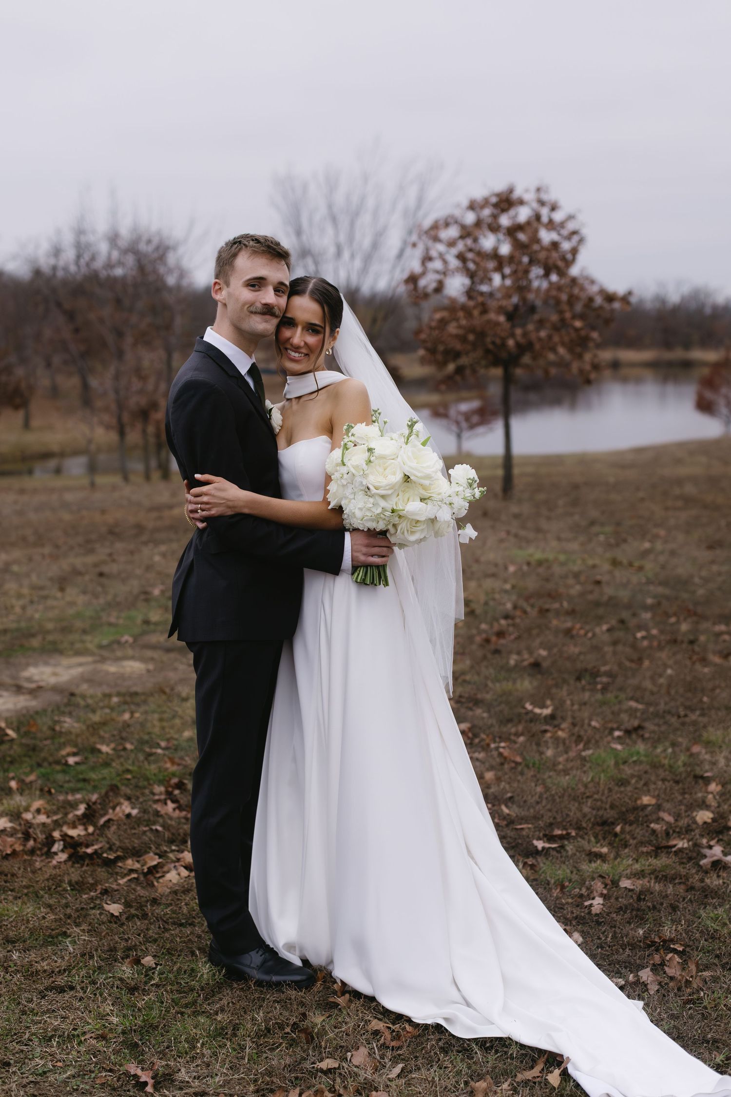 Wedding couple poses in autumn field with bride holding white bouquet and wearing elegant flowing gown.