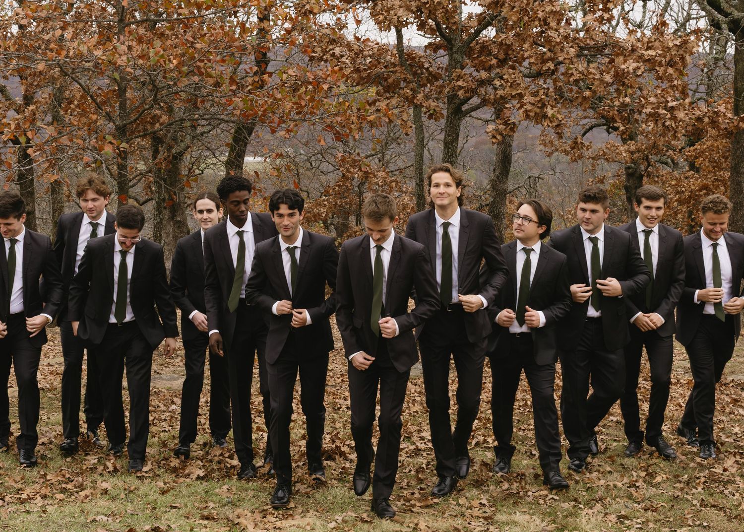 Groomsmen in black suits and ties stand in formation against autumn landscape.