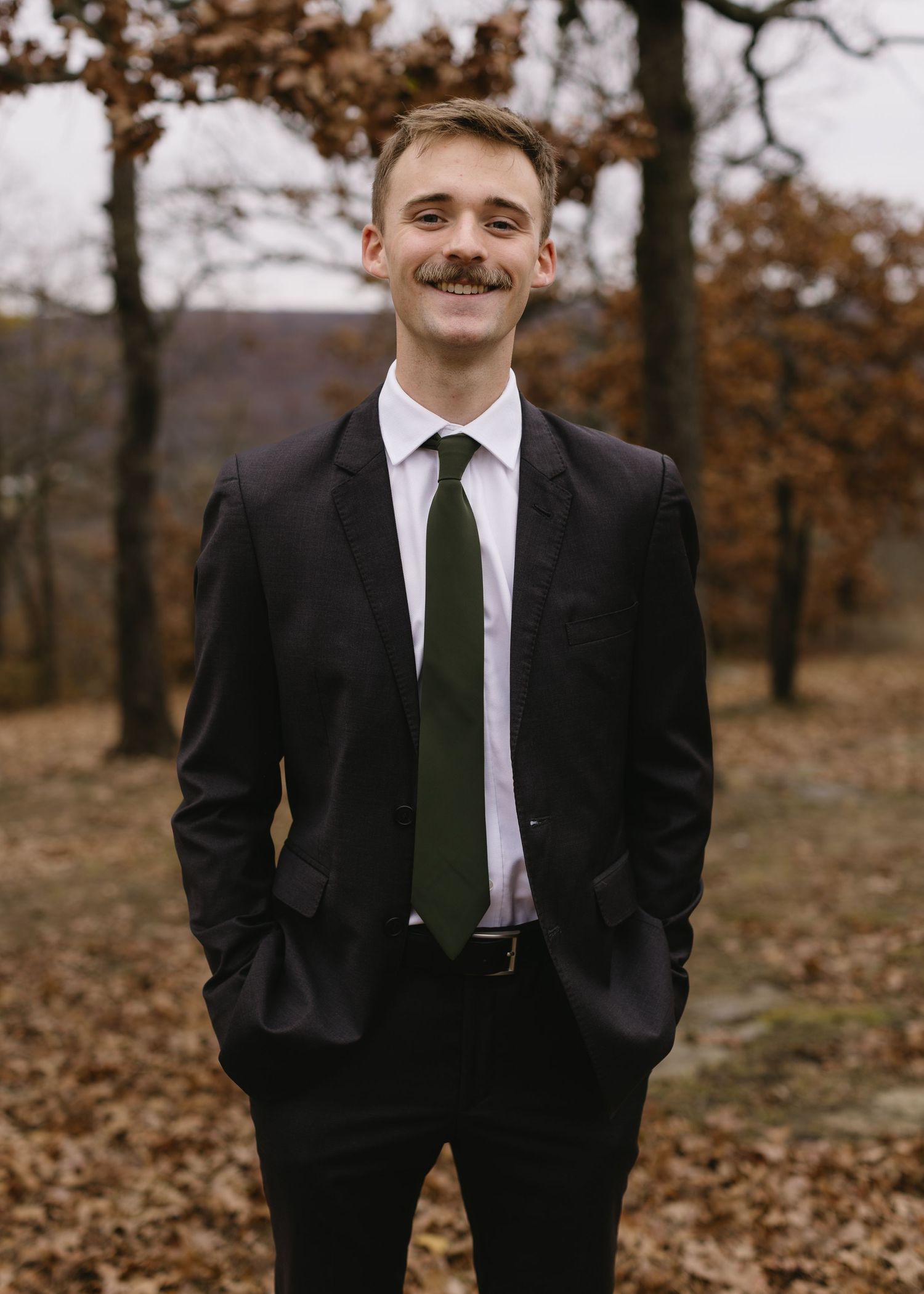 A suited figure stands in a fall landscape with autumn leaves and bare trees.