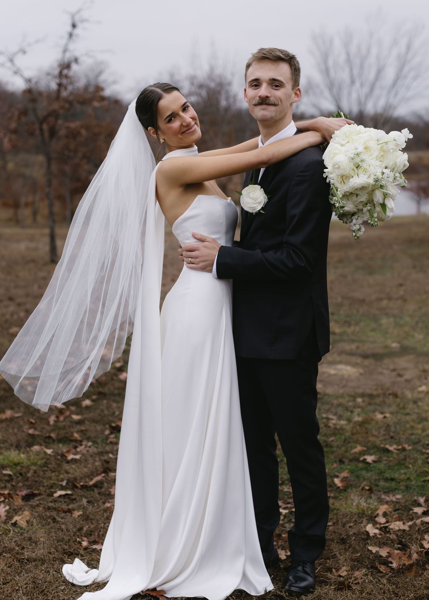 Couple embraces in outdoor fall setting with flowing veil and white floral bouquet.