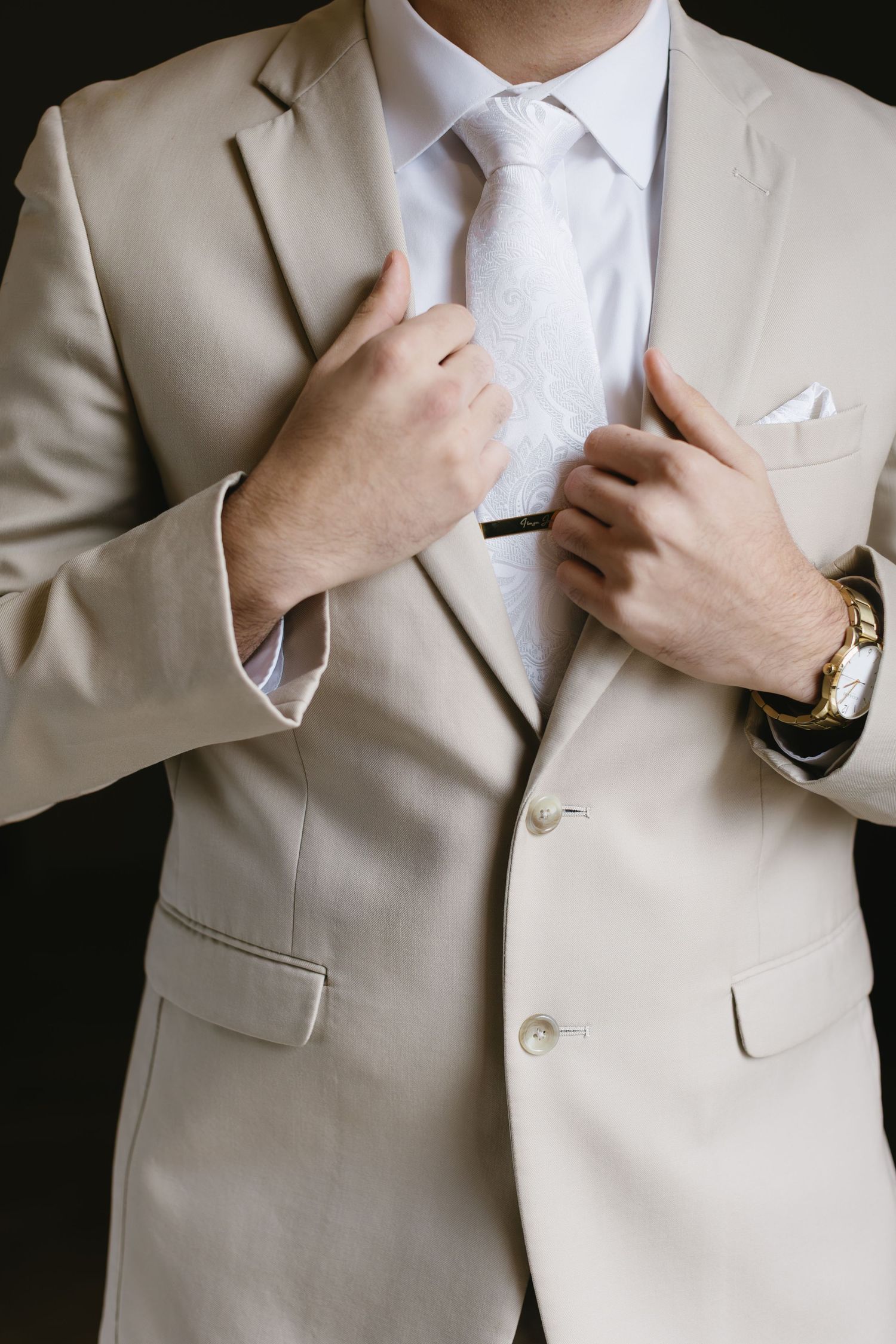 Close-up details of groom adjusting beige suit jacket and white shirt.