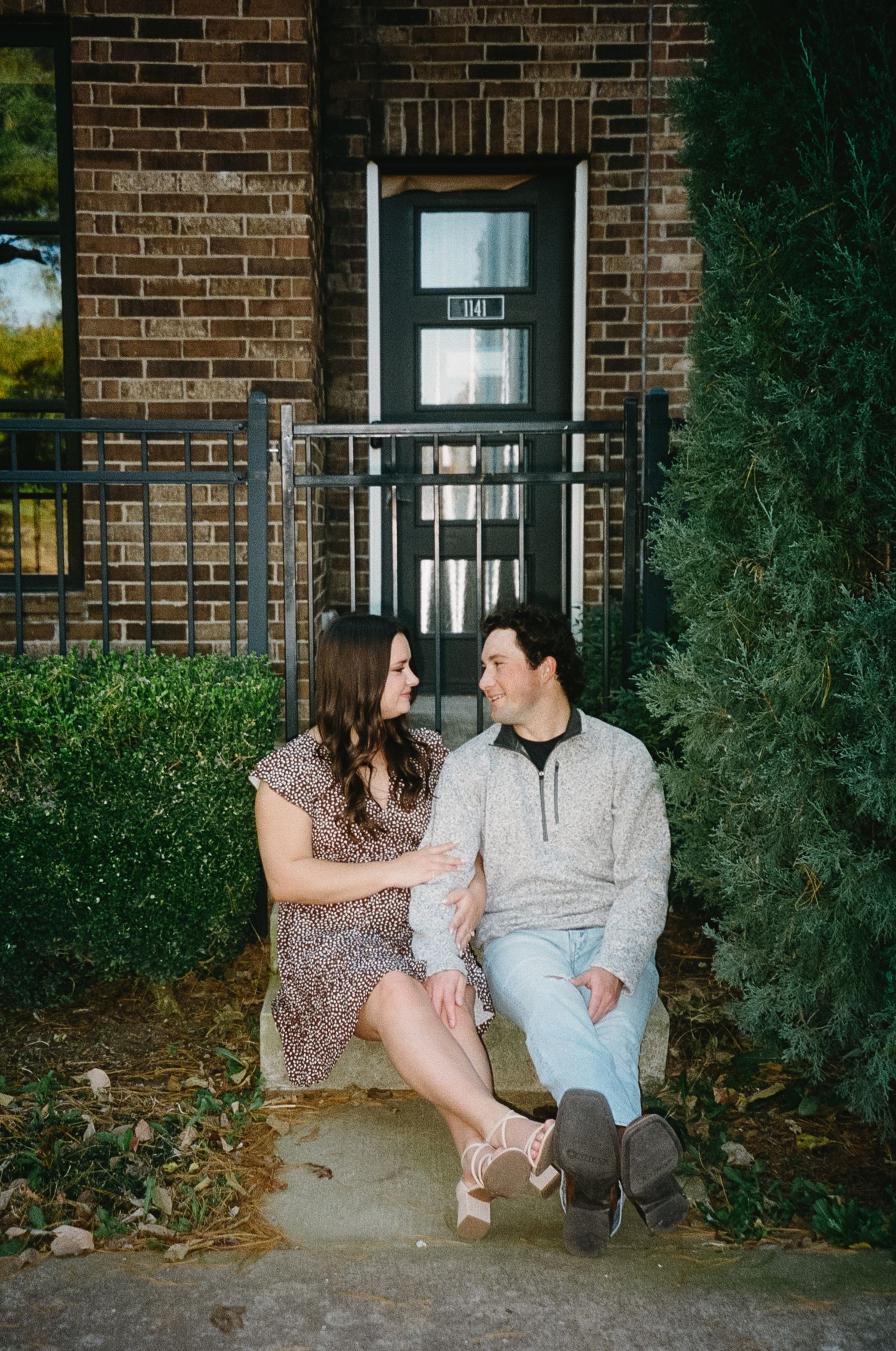 A couple sits together on a bench outside a brick building on an autumn evening.