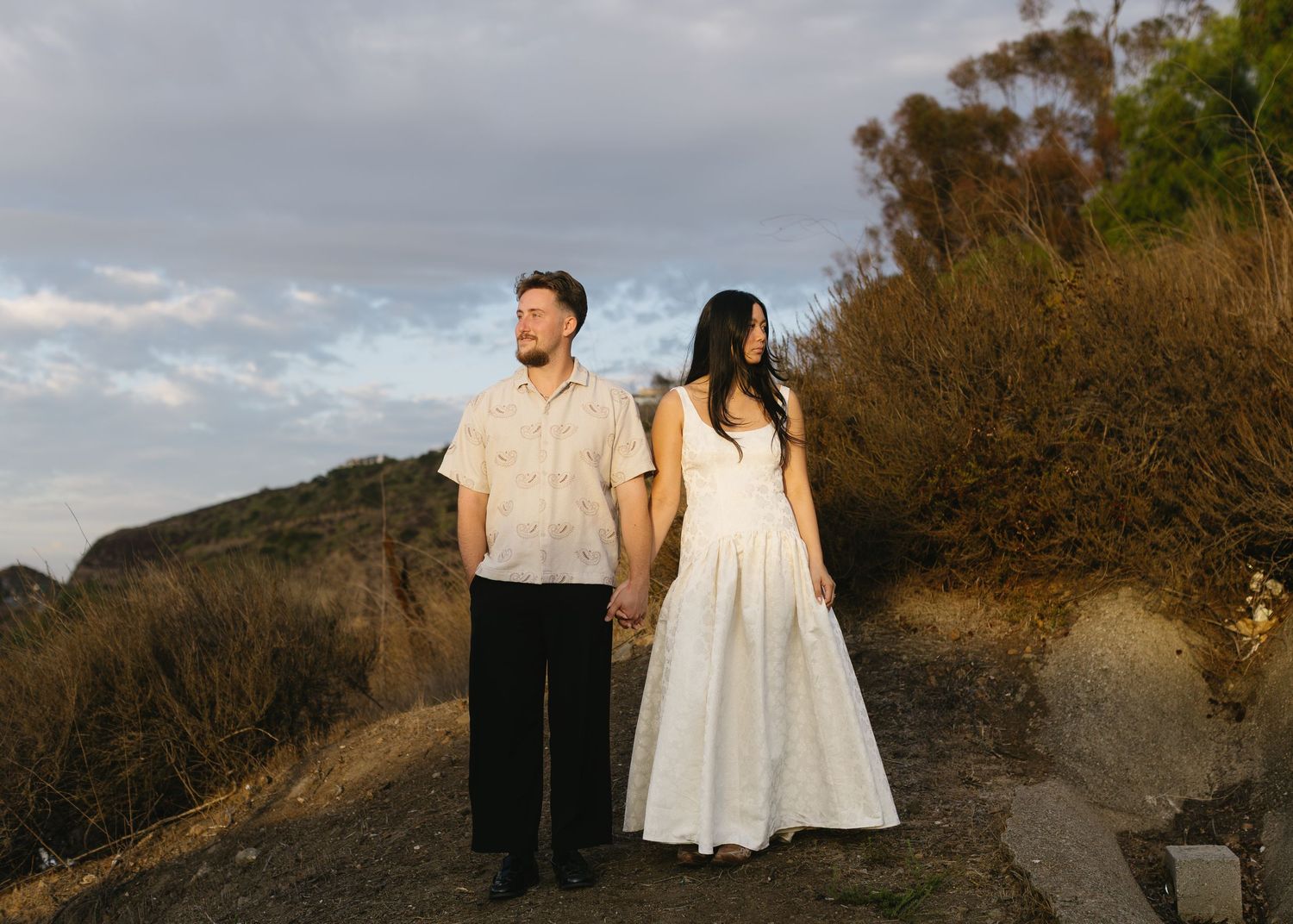 A couple in light-colored outfits walks along a scenic mountain path during golden hour.