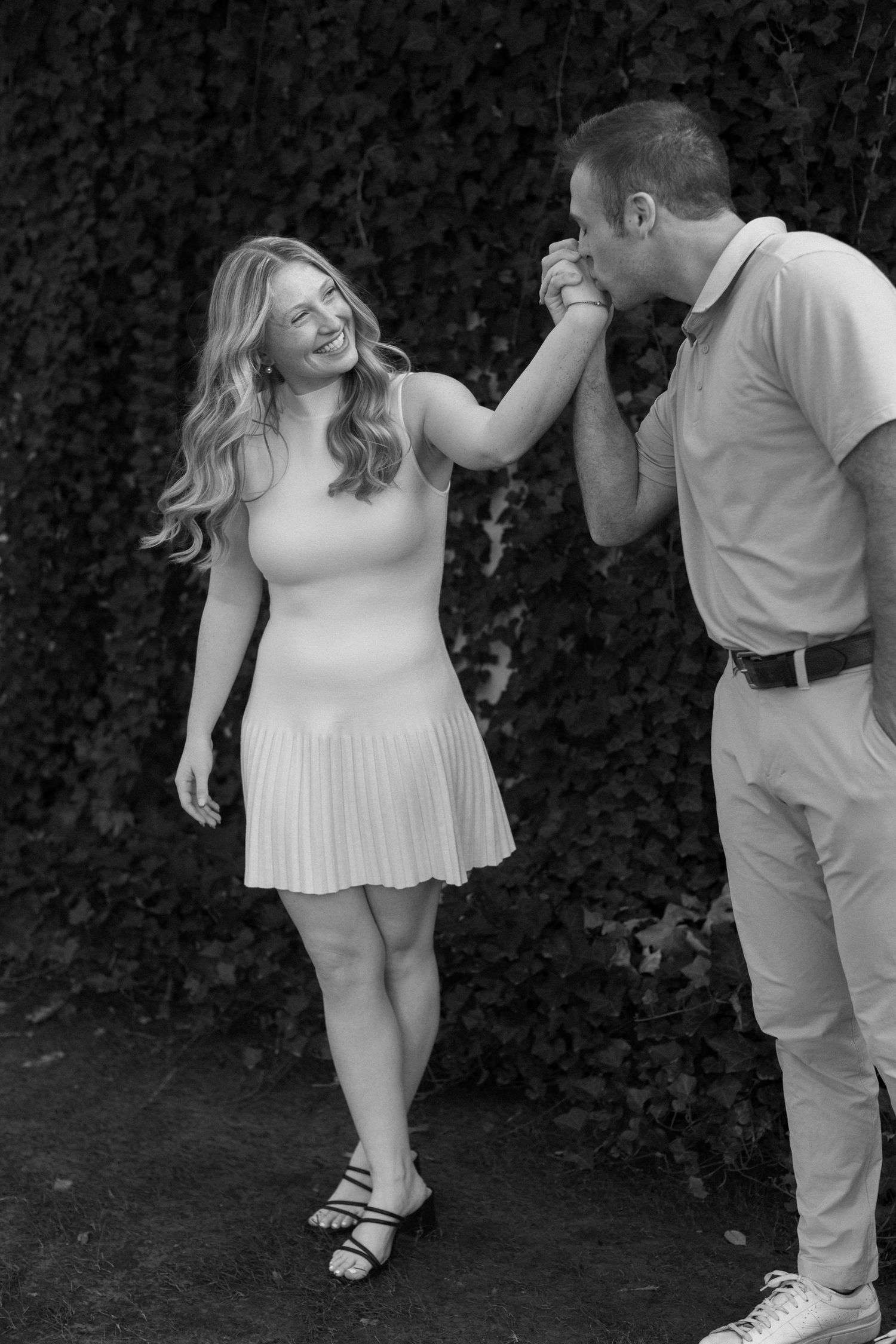 A couple in white clothing dance playfully together in a garden setting against a dark hedge background.