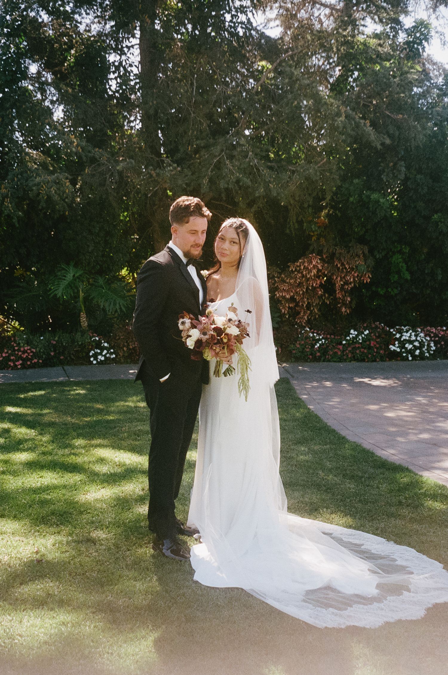 Newlyweds pose on a garden path with a flowing white wedding dress and veil.