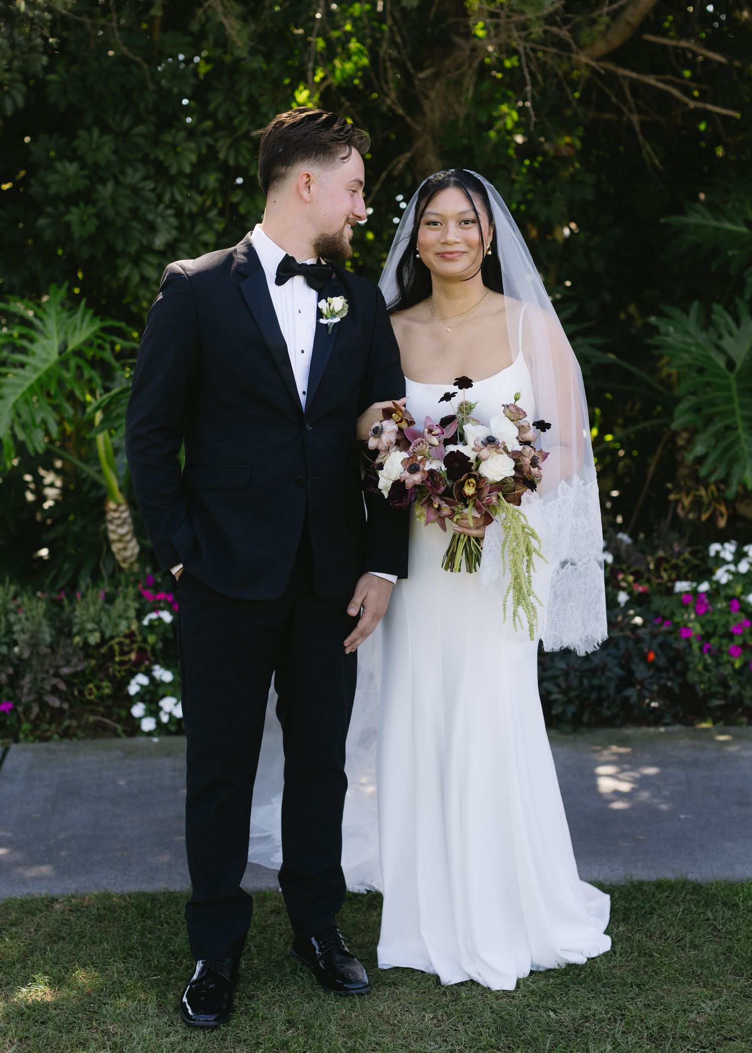 Newlywed couple poses in garden setting with beautiful floral bouquet and formal attire.